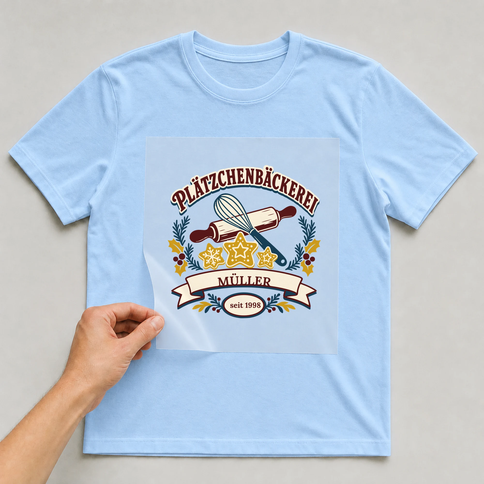 Bügelbild Plätzchenbäckerei mit Nachname und Jahr auf hellblauem T-Shirt, Folie wird aufgelegt