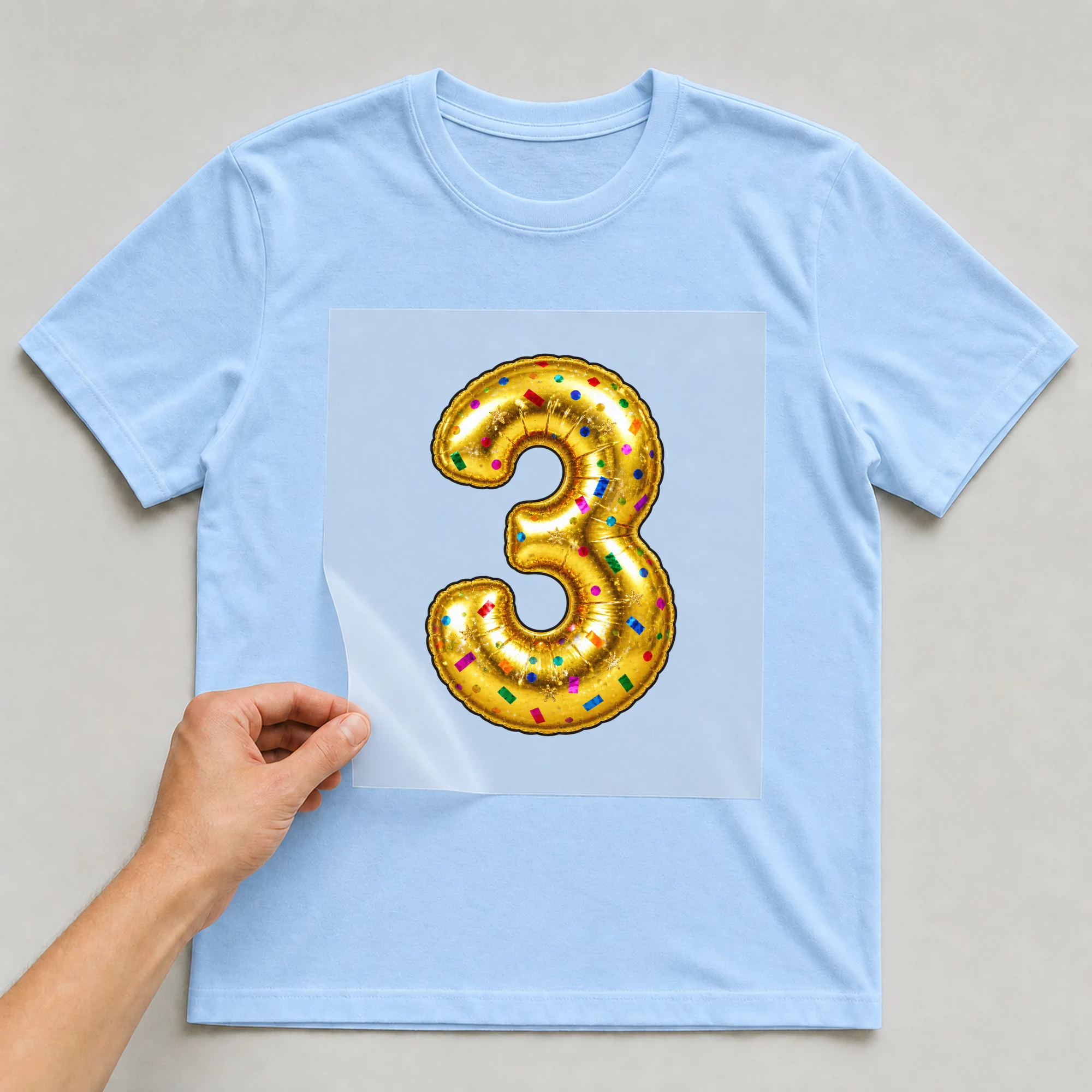 Bügelbild „Zahl 3 im Konfetti-Metallic-Look“ auf babyblauem T-Shirt, Folie wird aufgelegt