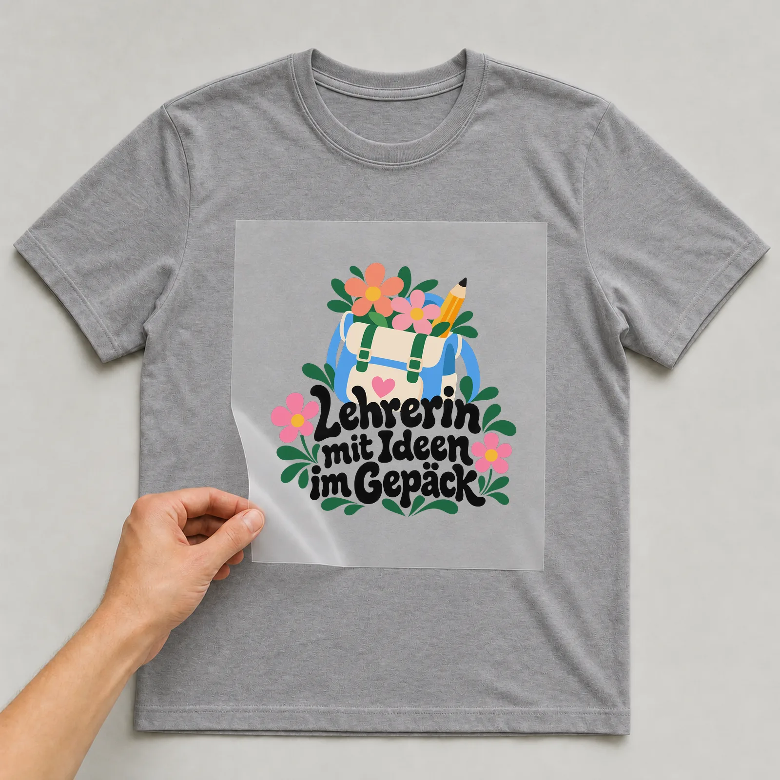 Bügelbild „Lehrerin mit Ideen im Gepäck“ auf grauem T-Shirt, Folie wird aufgelegt