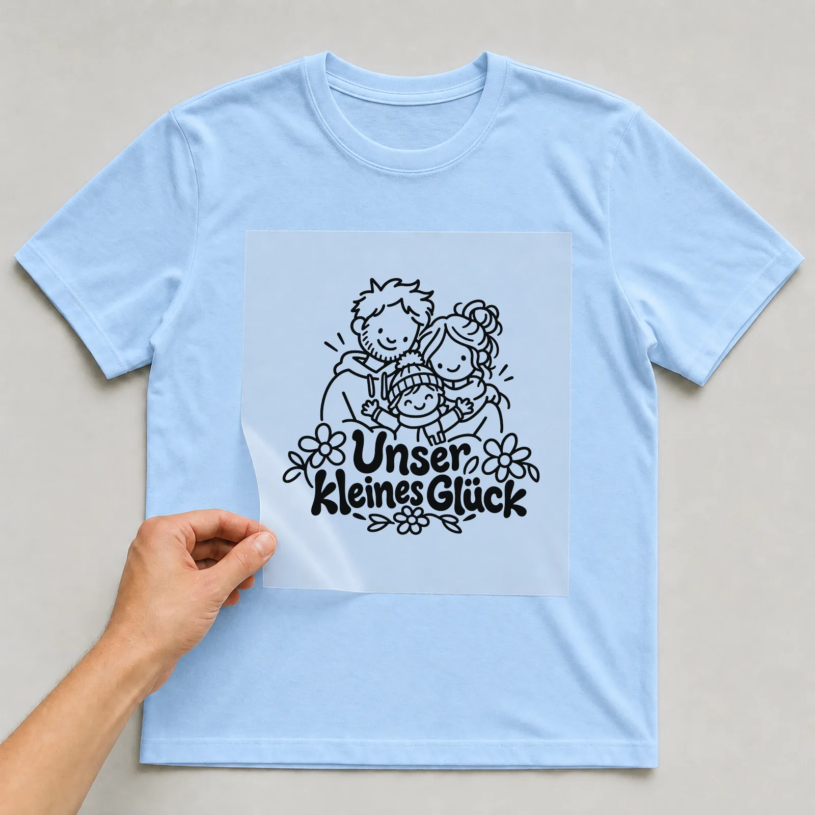 Bügelbild „Unser kleines Glück“ auf hellblauem T-Shirt, Folie wird aufgelegt
