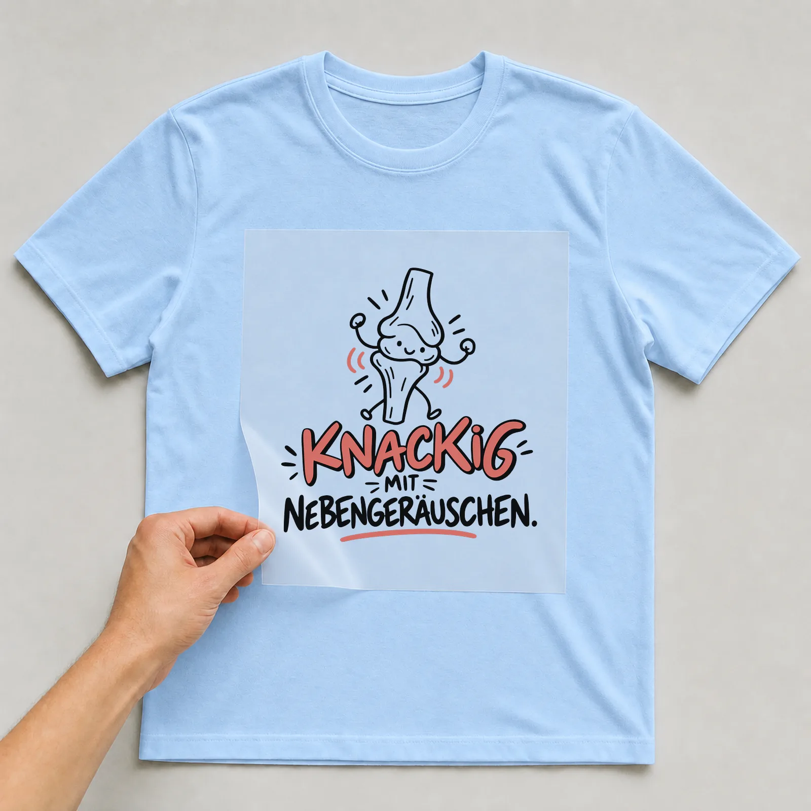 Bügelbild „Knackig mit Nebengeräuschen“ auf hellblauem T-Shirt, Folie wird aufgelegt