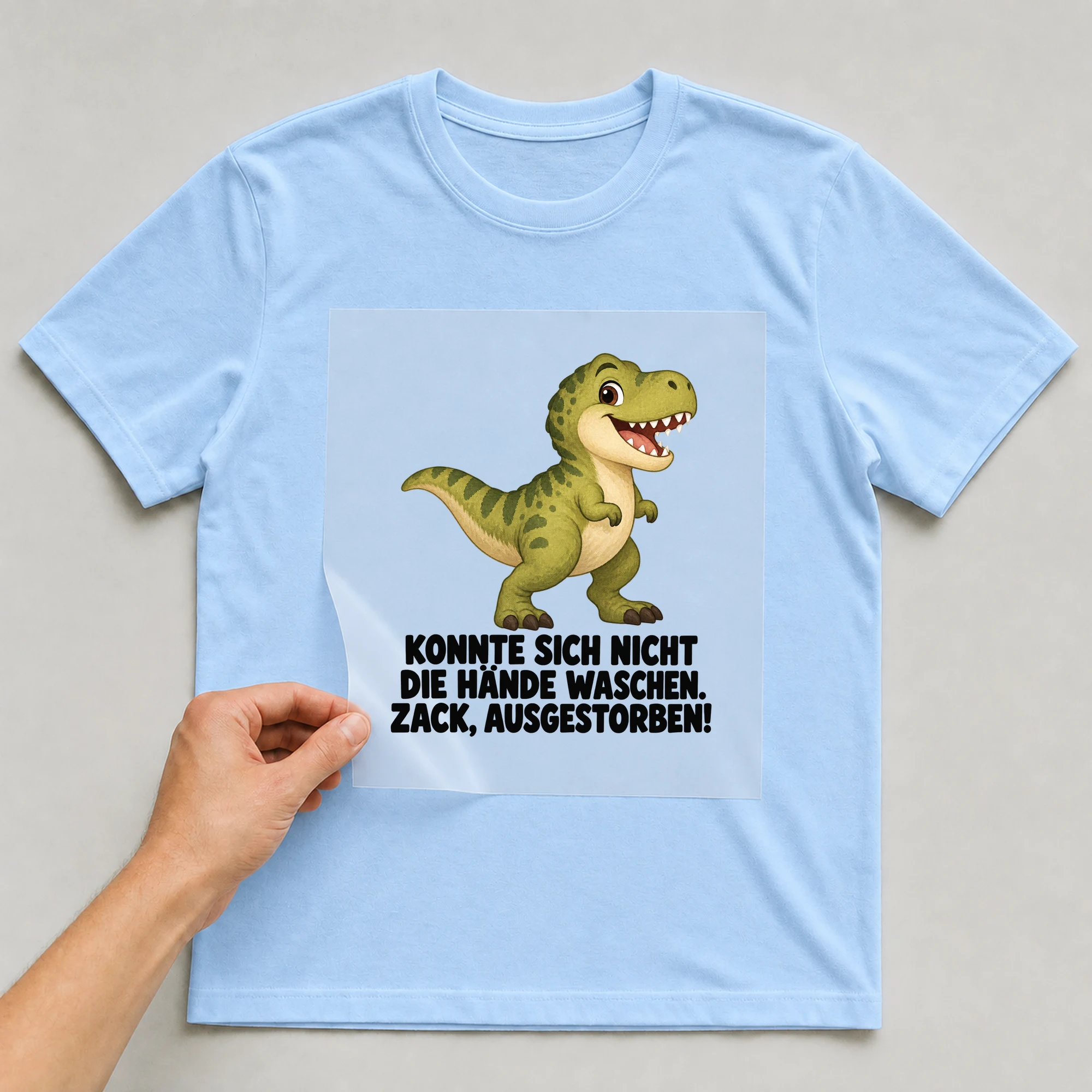 Bügelbild „Konnte Sich Nicht Die Hände Waschen. Zack, Ausgestorben!“ auf babyblauem T-Shirt, Folie wird aufgelegt