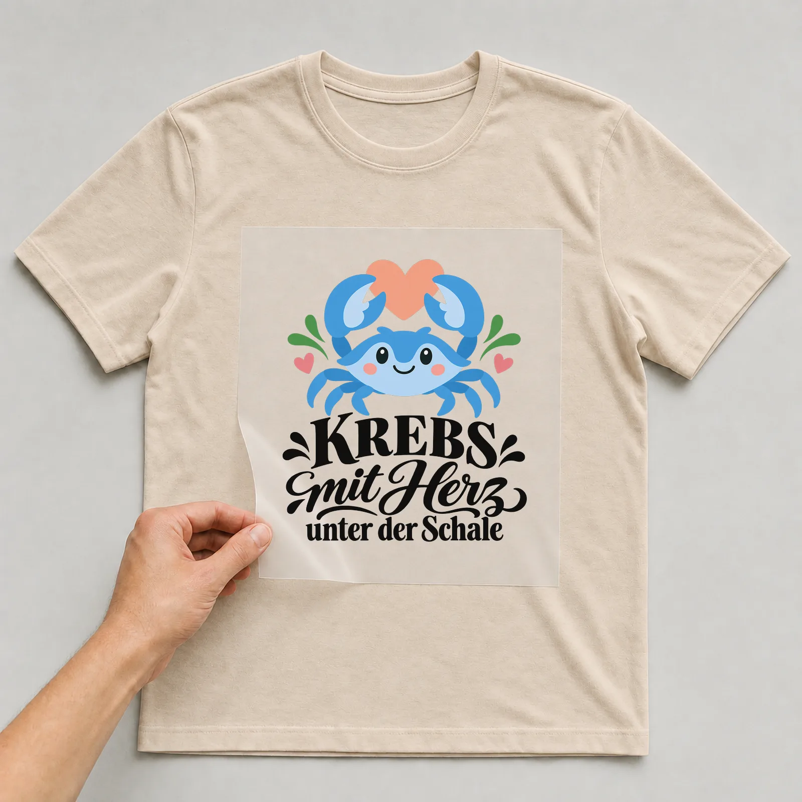 Bügelbild „KREBS mit Herz unter der Schale“ auf beigem T-Shirt, Folie wird aufgelegt