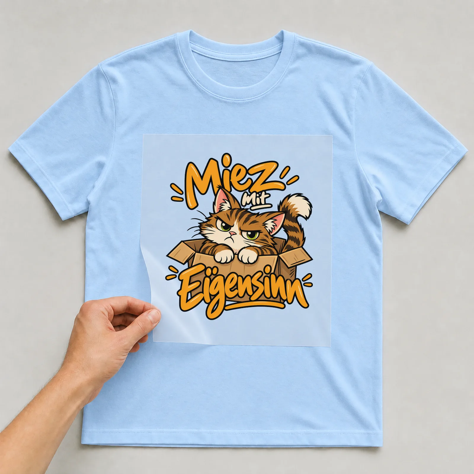 Bügelbild „Miez mit Eigensinn“ auf hellblauem T-Shirt, Folie wird aufgelegt