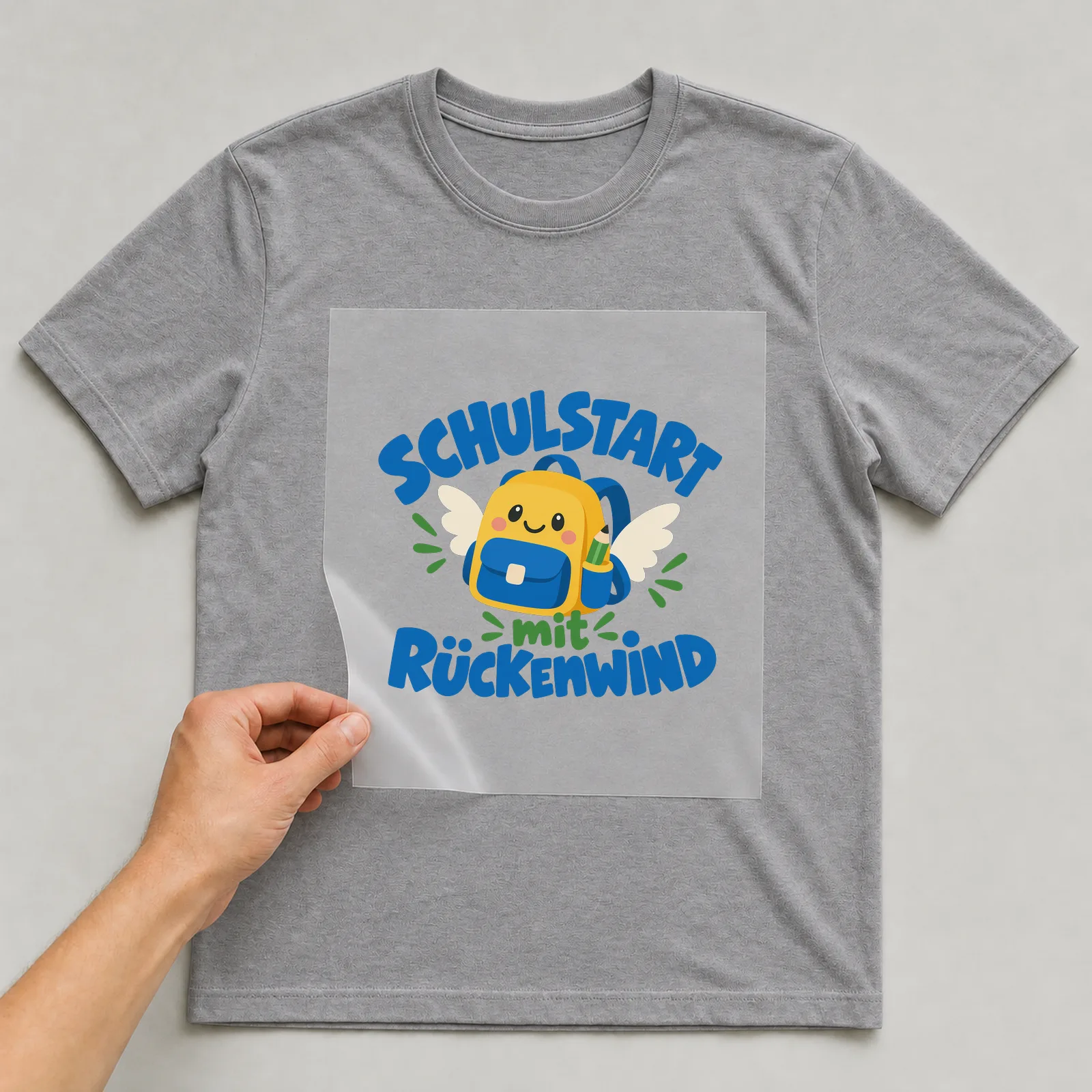 Bügelbild „Schulstart mit Rückenwind“ auf grauem T-Shirt, Folie wird aufgelegt