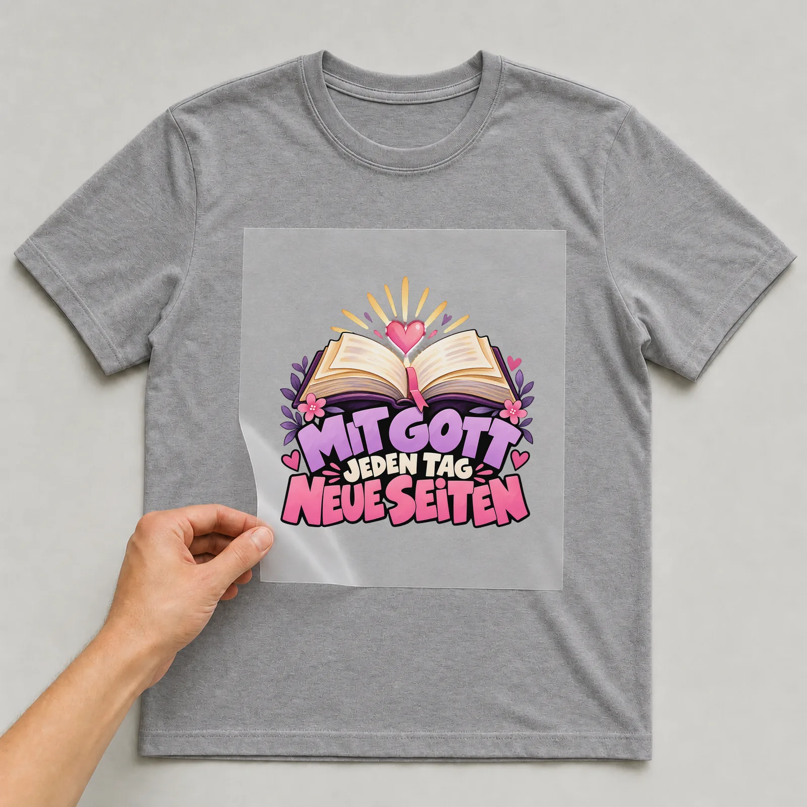 Bügelbild „Mit Gott jeden Tag neue Seiten“ auf grauem T-Shirt, Folie wird aufgelegt