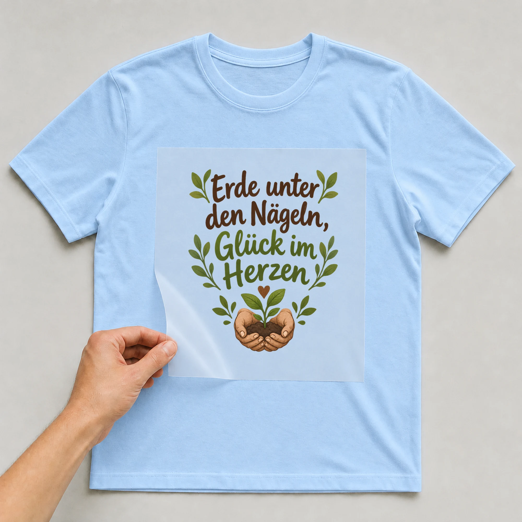 Bügelbild „Erde unter den naegeln glueck im“ auf babyblauem T-Shirt, Folie wird aufgelegt