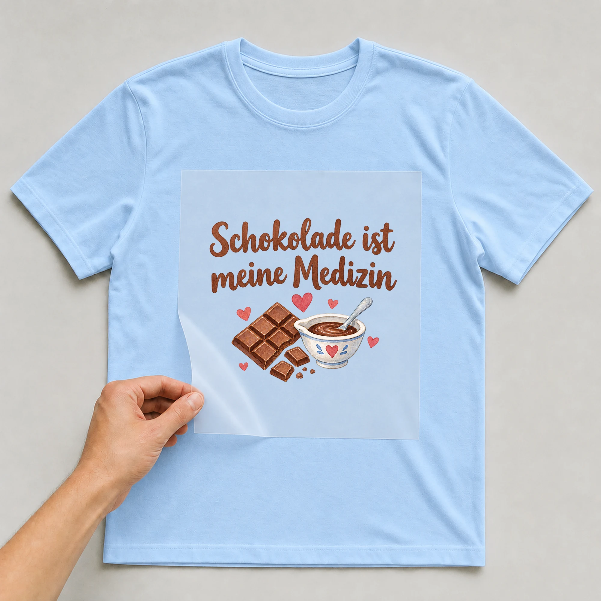 Bügelbild „Schokolade ist meine Medizin“ auf babyblauem T-Shirt, Folie wird aufgelegt