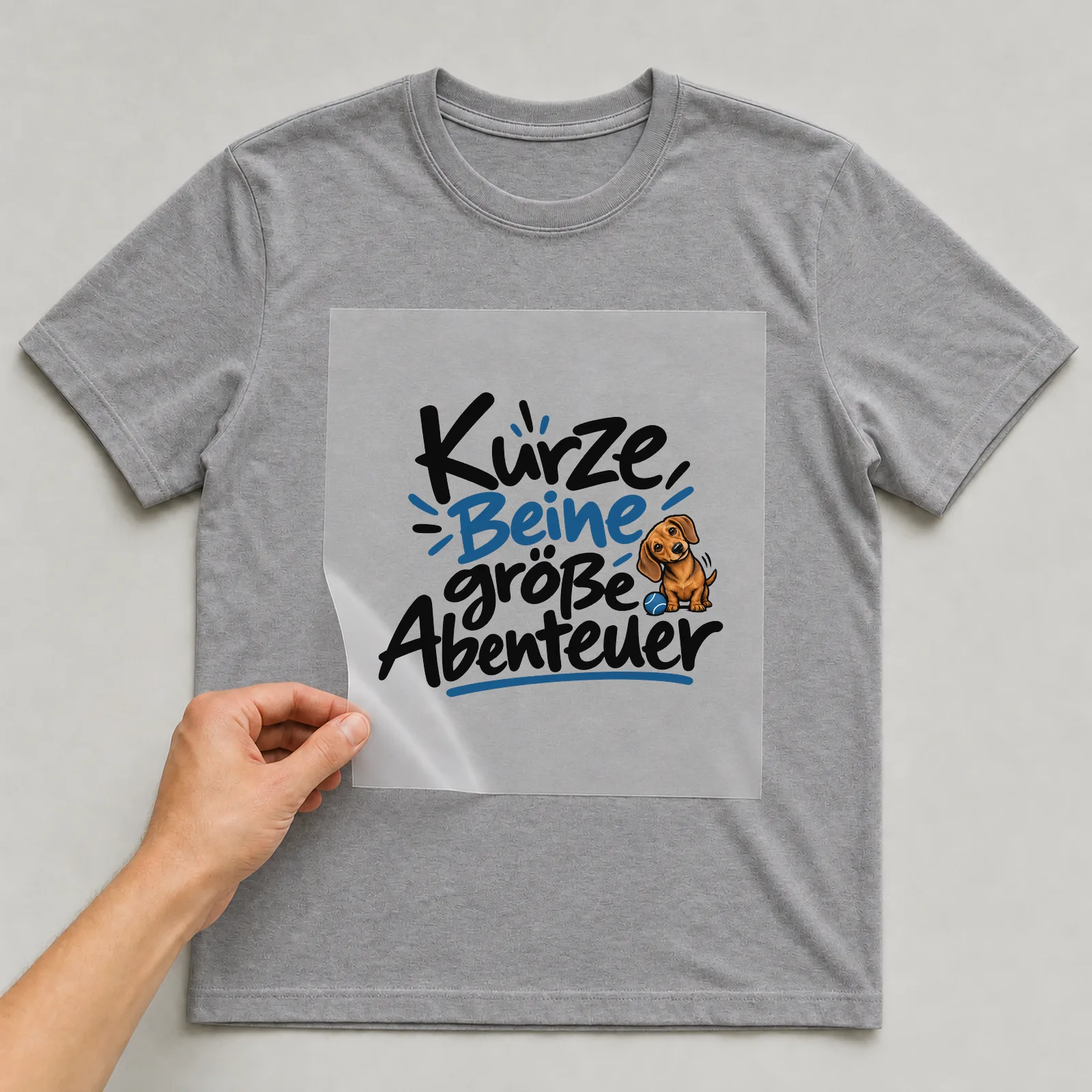 Bügelbild „Kurze Beine große Abenteuer“ auf grauem T-Shirt, Folie wird aufgelegt