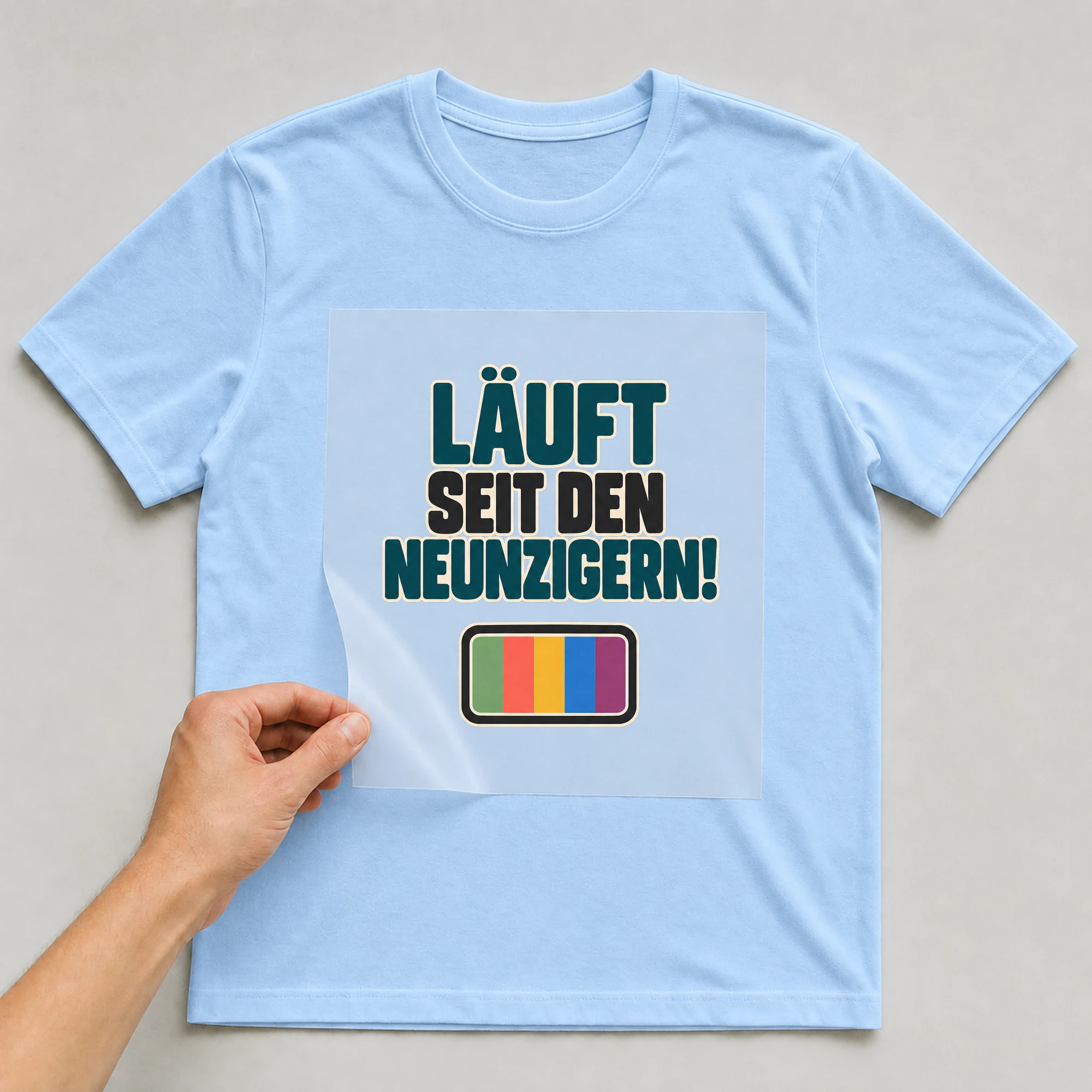 Bügelbild „LÄUFT SEIT DEN NEUNZIGERN!“ auf babyblauem T-Shirt, Folie wird aufgelegt