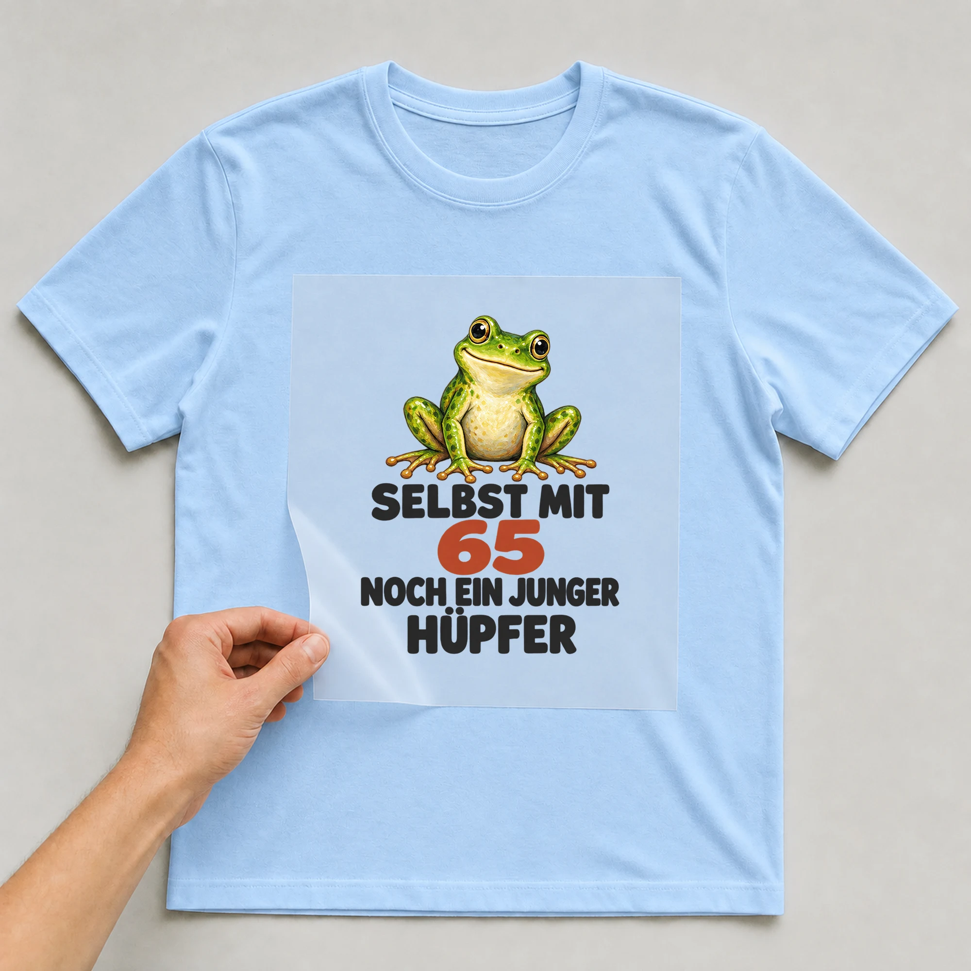 Bügelbild „Selbst mit 65 noch ein junger Hüpfer“ auf babyblauem T-Shirt, Folie wird aufgelegt
