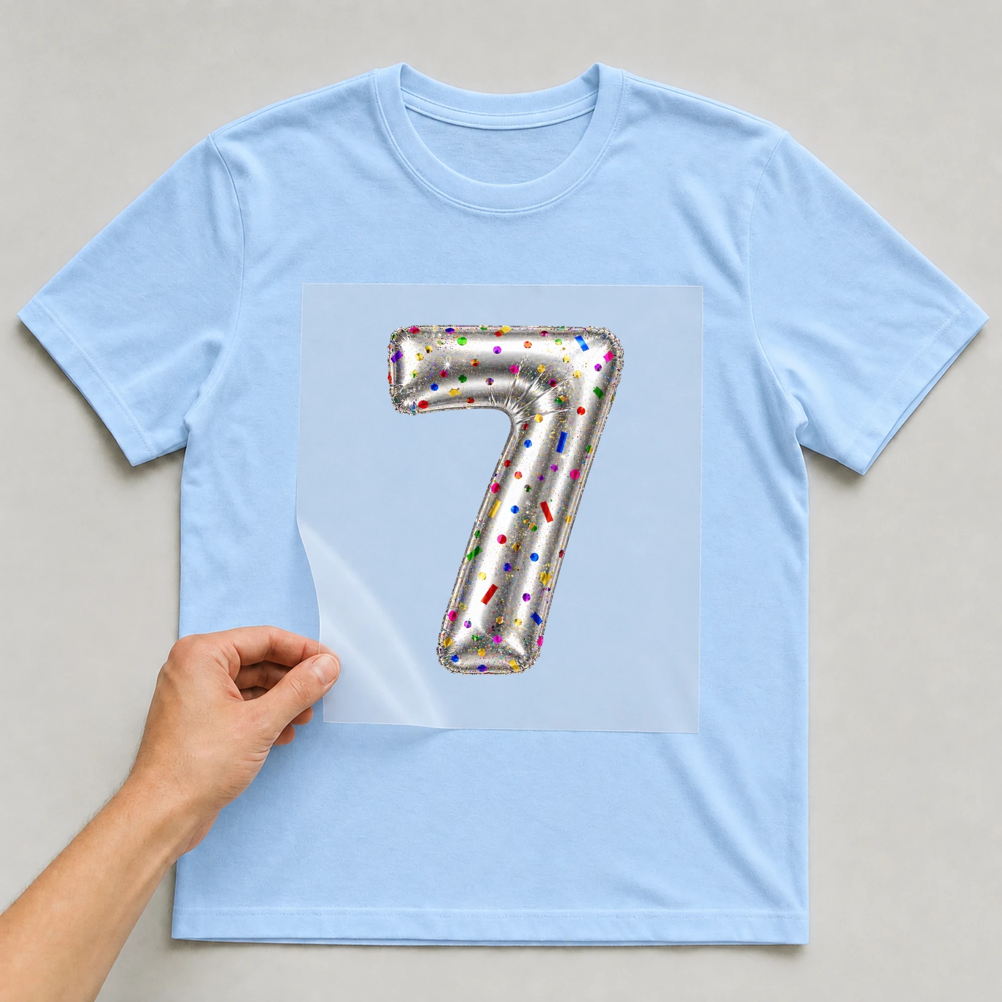 Bügelbild „Zahl 7 im Konfetti-Metallic-Look“ auf babyblauem T-Shirt, Folie wird aufgelegt
