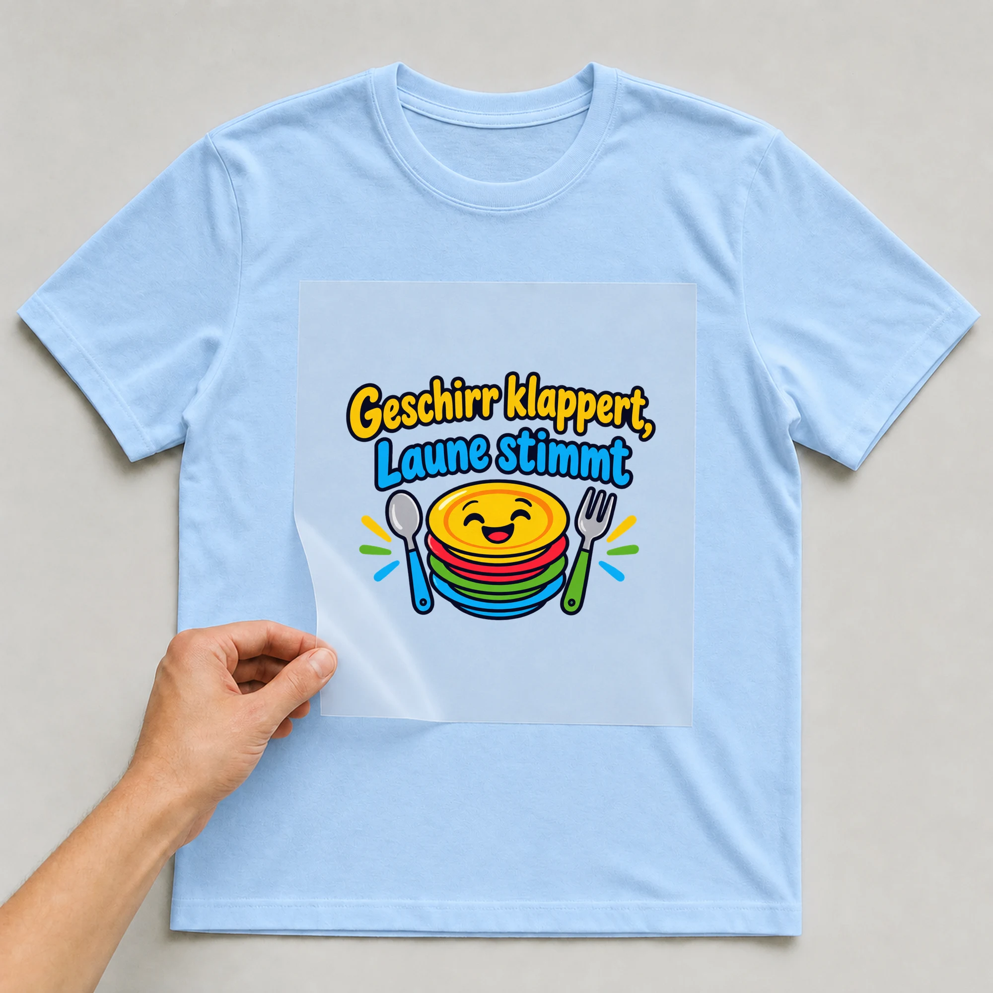 Bügelbild „Geschirr klappert, Laune stimmt“ auf babyblauem T-Shirt, Folie wird aufgelegt