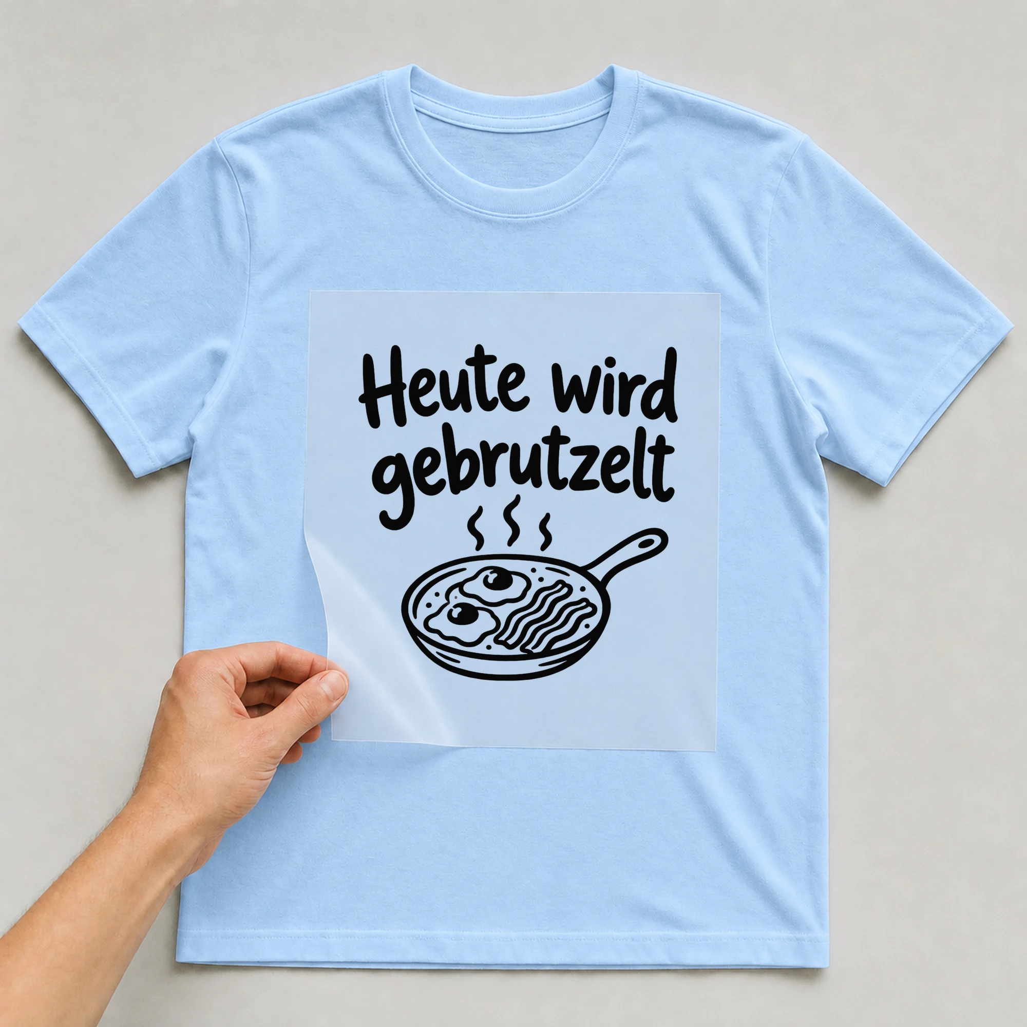 Bügelbild „Heute wird gebrutzelt“ auf babyblauem T-Shirt, Folie wird aufgelegt
