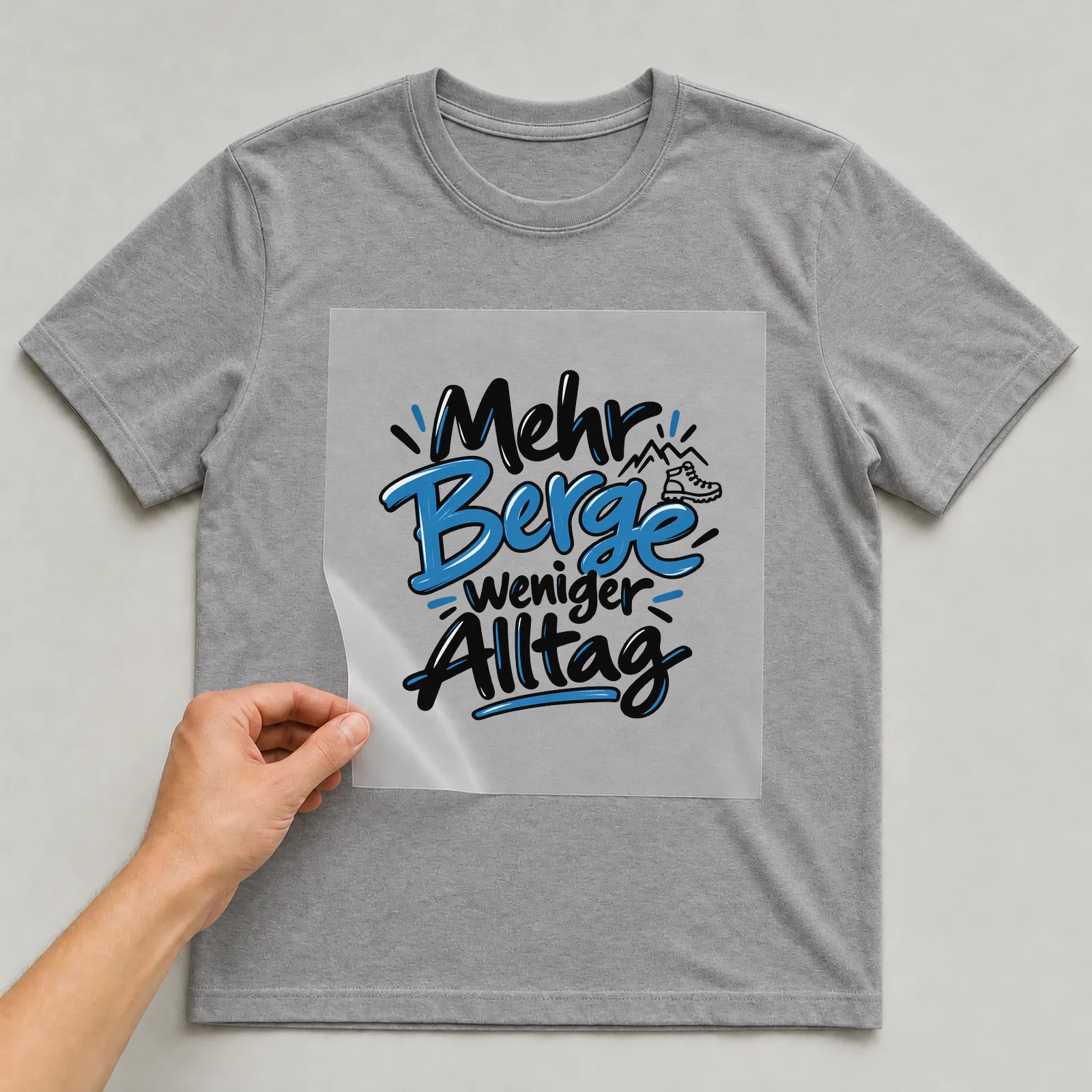 Bügelbild „Mehr Berge weniger Alltag“ auf grauem T-Shirt, Folie wird aufgelegt