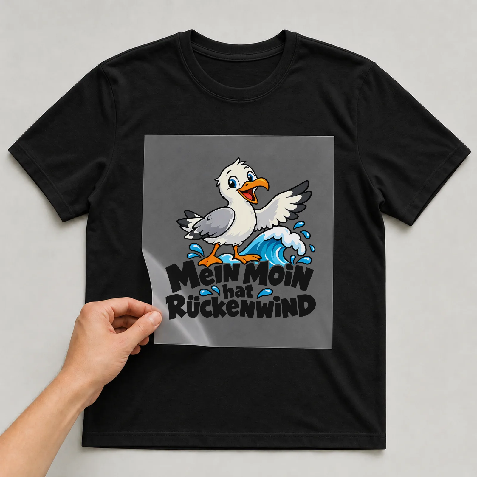 Bügelbild „MEIN MOIN hat RÜCKENWIND“ auf schwarzem T-Shirt, Folie wird aufgelegt