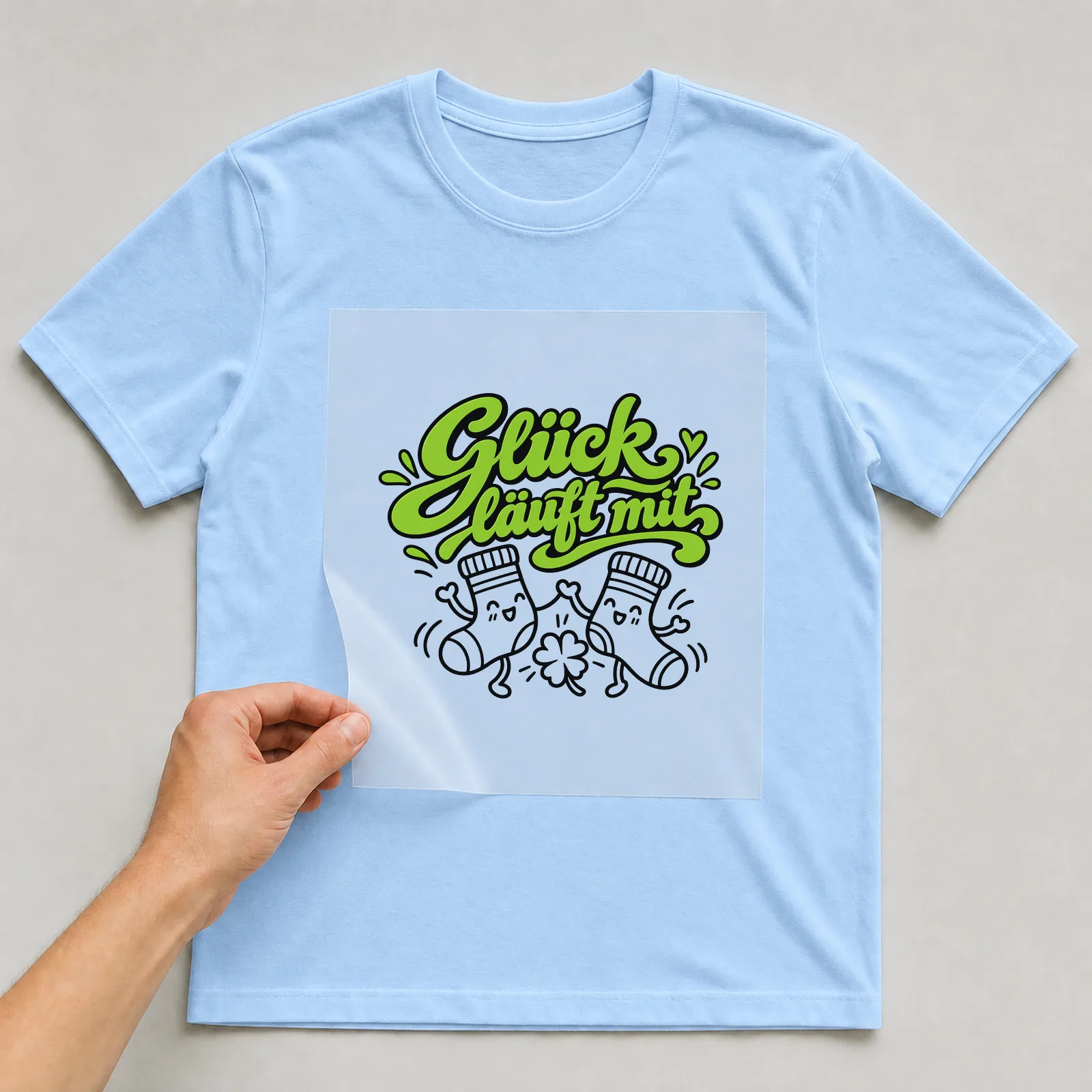 Bügelbild „Glück läuft mit“ auf hellblauem T-Shirt, Folie wird aufgelegt