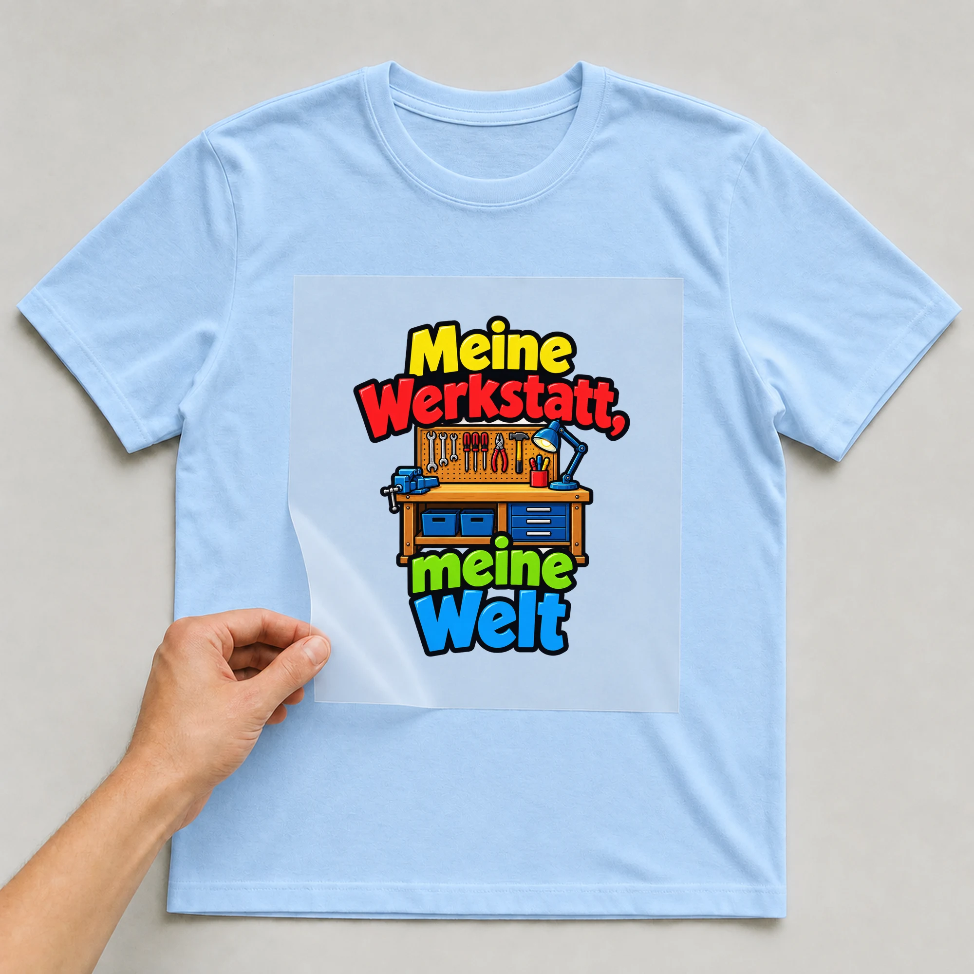 Bügelbild „Meine Werkstatt, meine Welt“ auf babyblauem T-Shirt, Folie wird aufgelegt