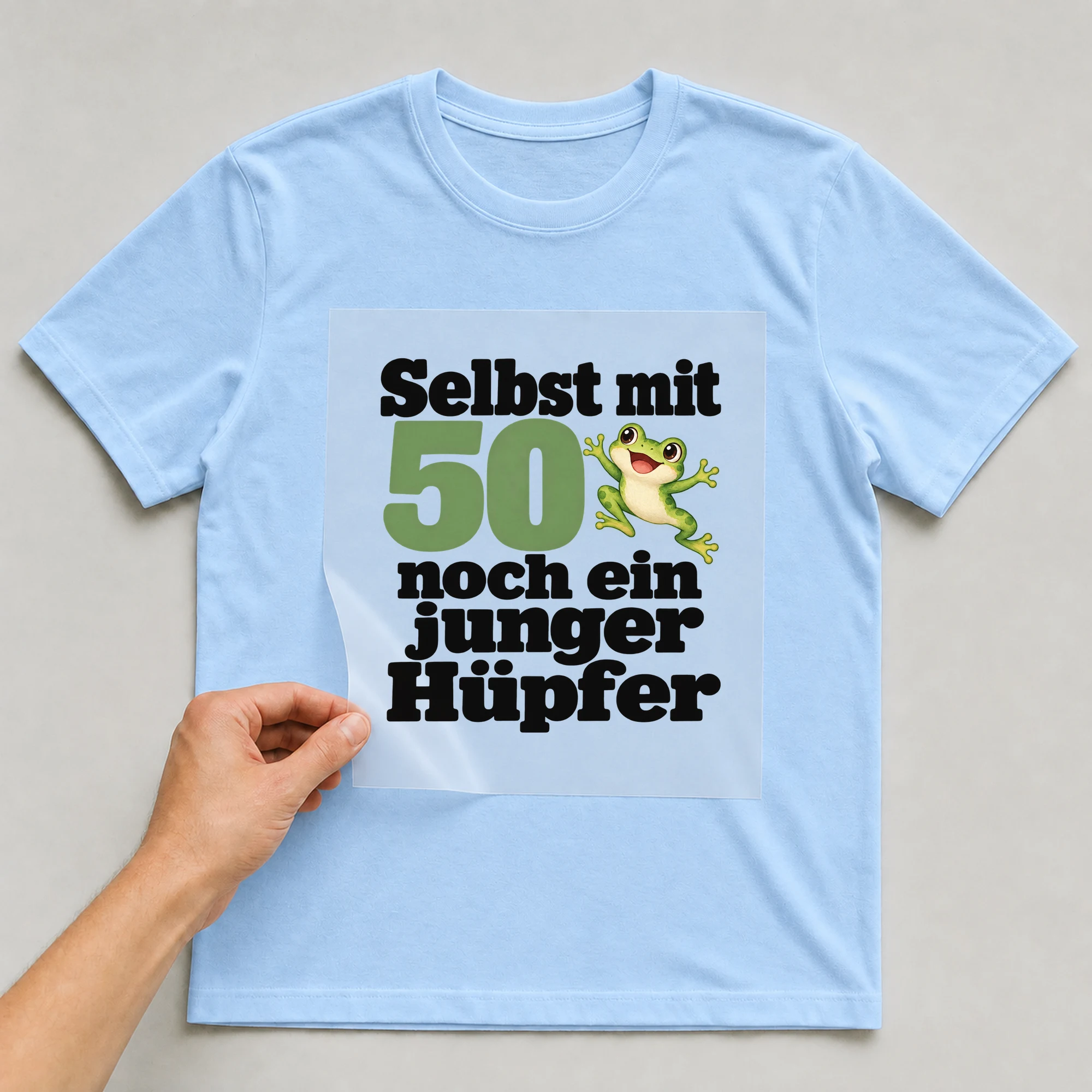 Bügelbild „Selbst mit 50 noch ein junger Hüpfer“ auf babyblauem T-Shirt, Folie wird aufgelegt