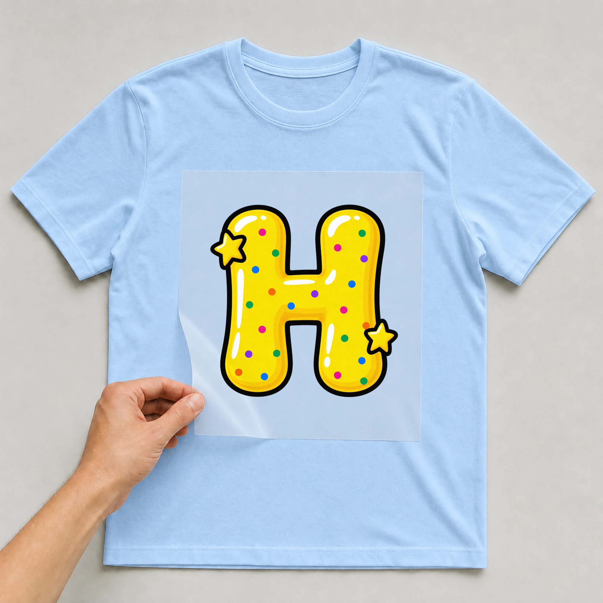 Bügelbild „Buchstabe H im Comic-Ballon-Stil“ auf babyblauem T-Shirt, Folie wird aufgelegt