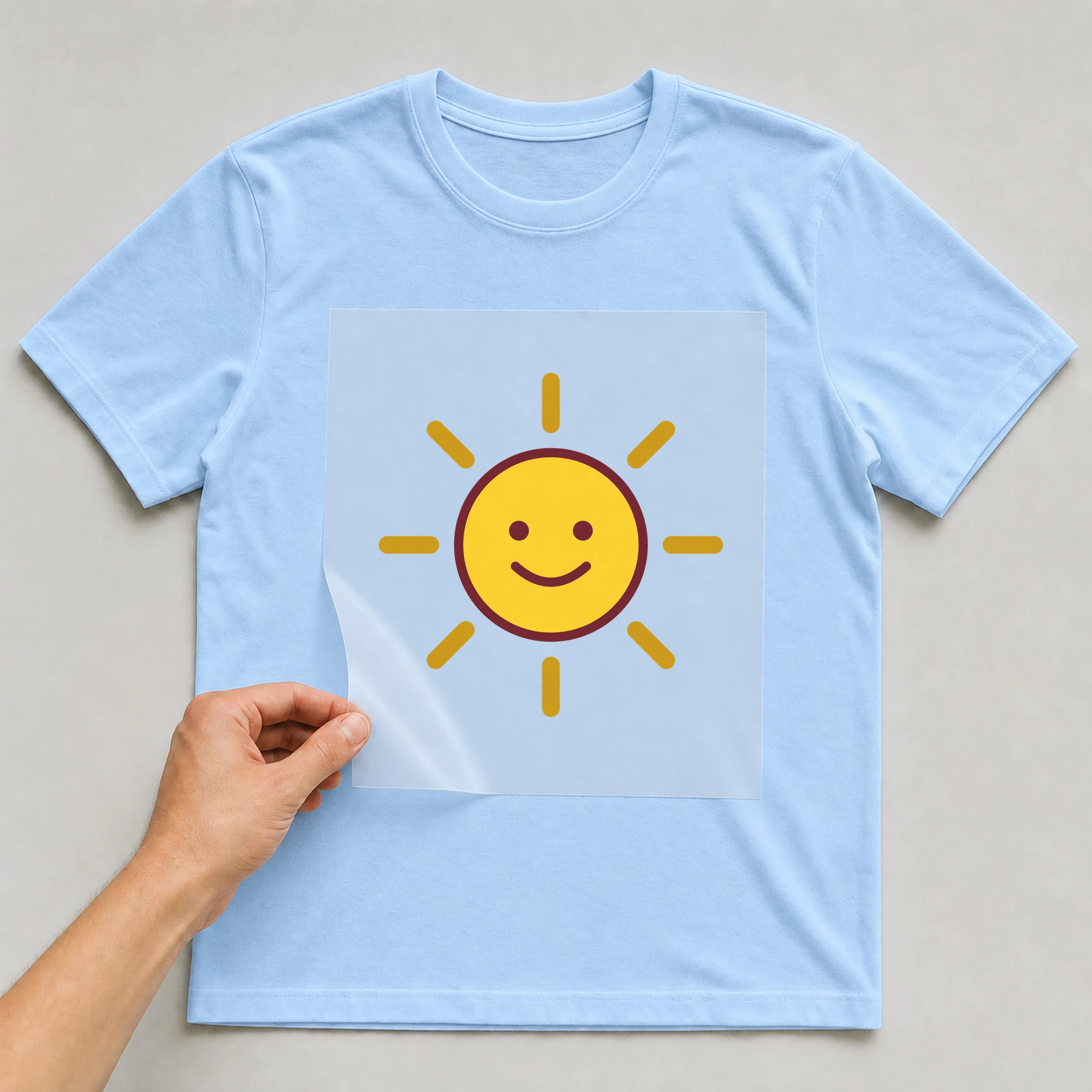 Bügelbild „Sonne mit Gesicht“ auf babyblauem T-Shirt, Folie wird aufgelegt