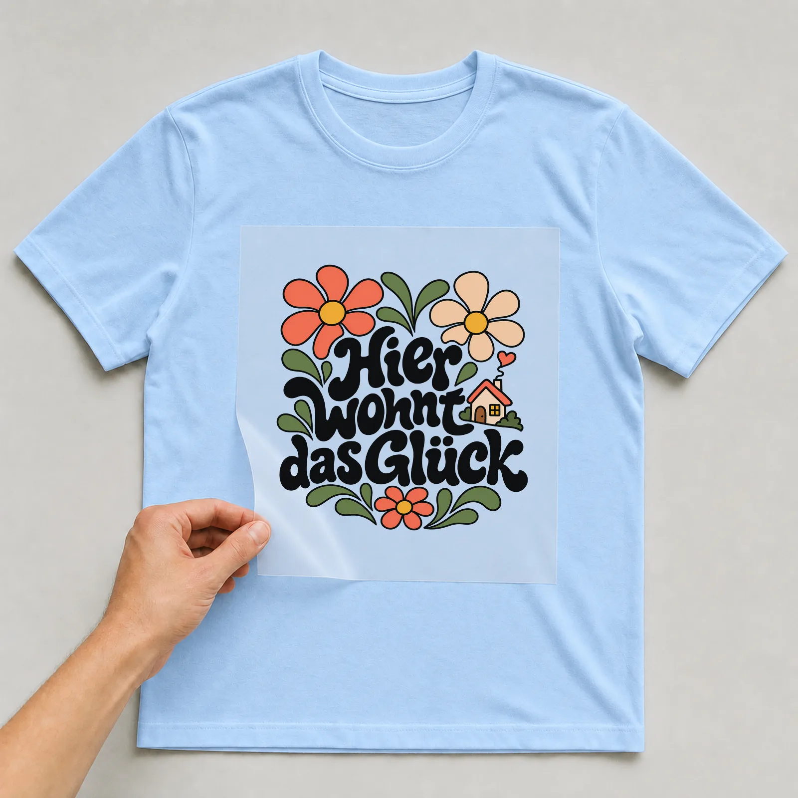 Bügelbild „Hier wohnt das Glück“ (Blumenlettering) auf hellblauem T-Shirt, Folie wird aufgelegt