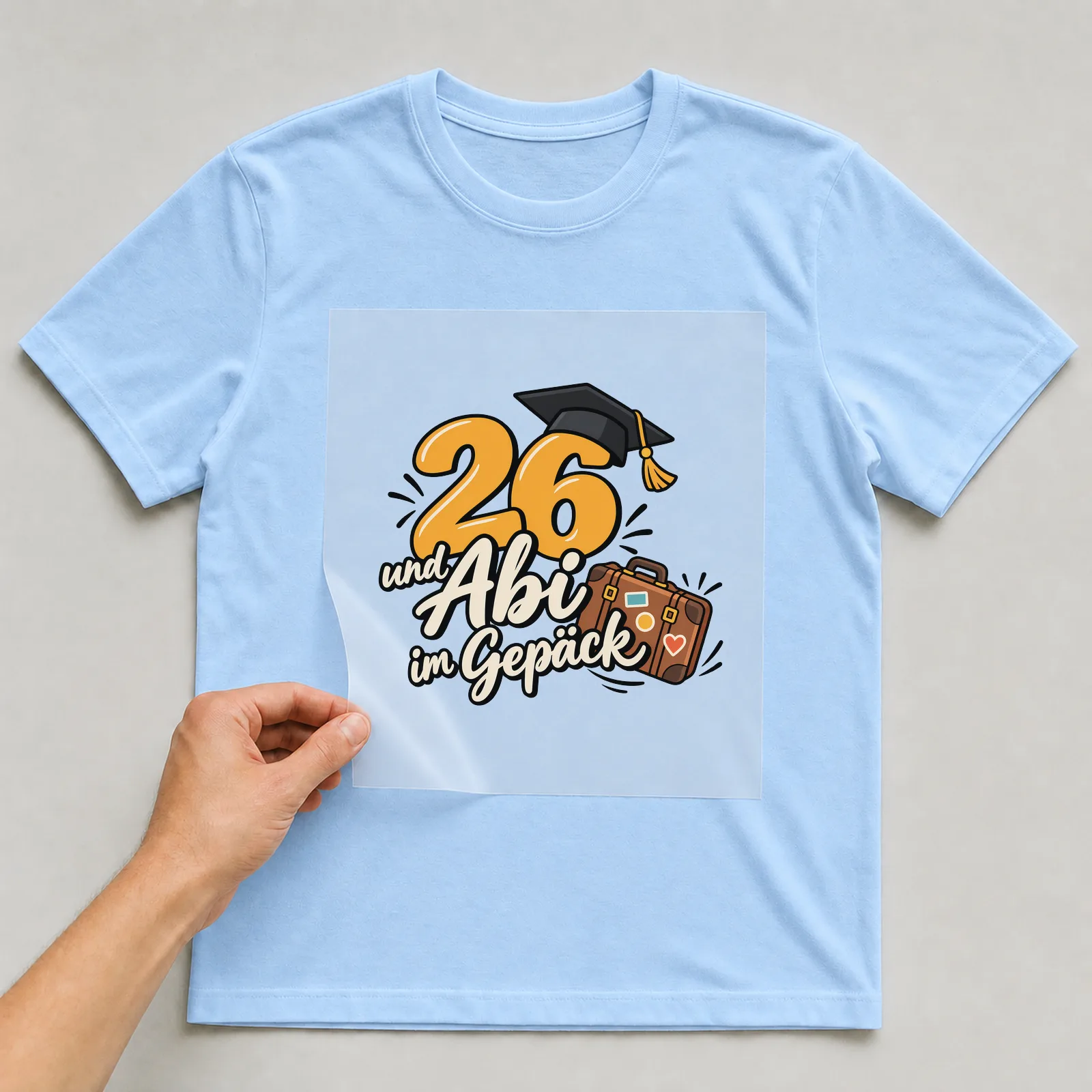 Bügelbild „26 und Abi im Gepäck“ auf hellblauem T-Shirt, Folie wird aufgelegt