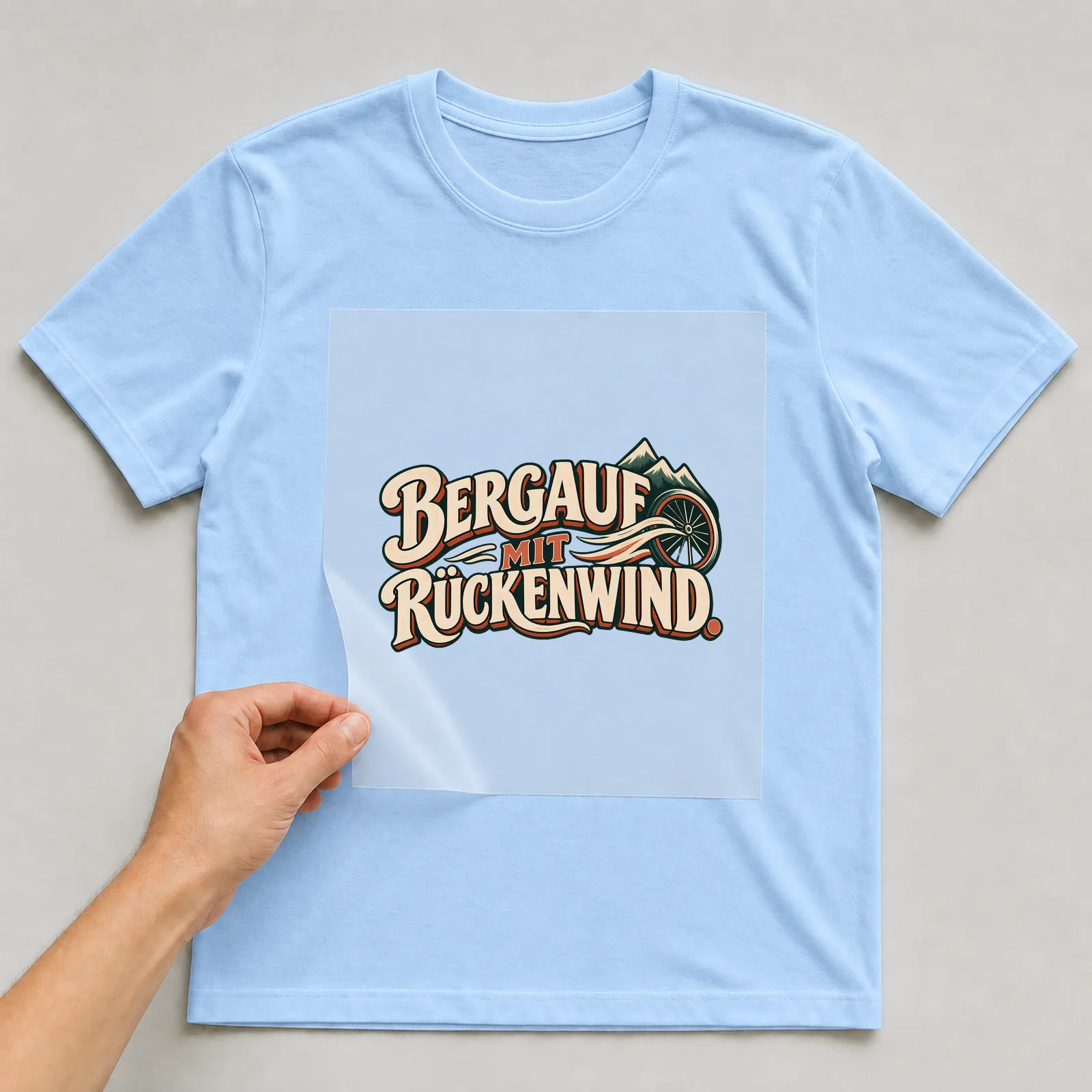 Bügelbild „Bergauf mit Rückenwind“ auf hellblauem T-Shirt, Folie wird aufgelegt
