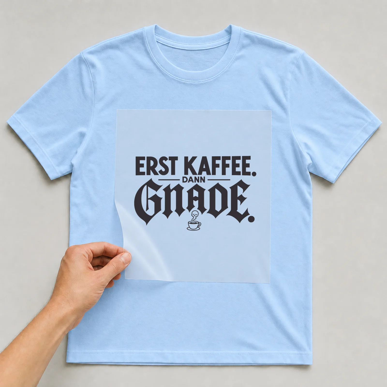 Bügelbild „Erst Kaffee. Dann Gnade.“ (Schrift Konturen) auf hellblauem T-Shirt, Folie wird aufgelegt