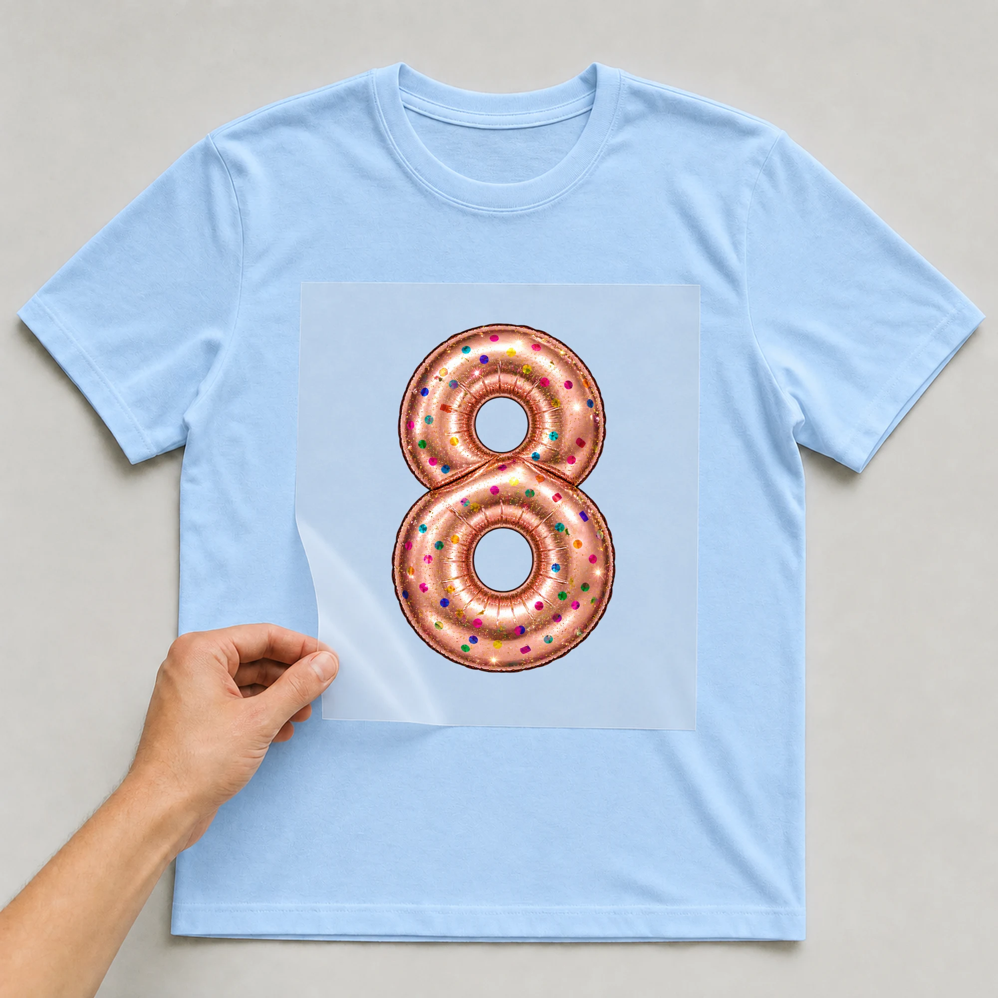 Bügelbild „Zahl 8 im Konfetti-Metallic-Look“ auf babyblauem T-Shirt, Folie wird aufgelegt