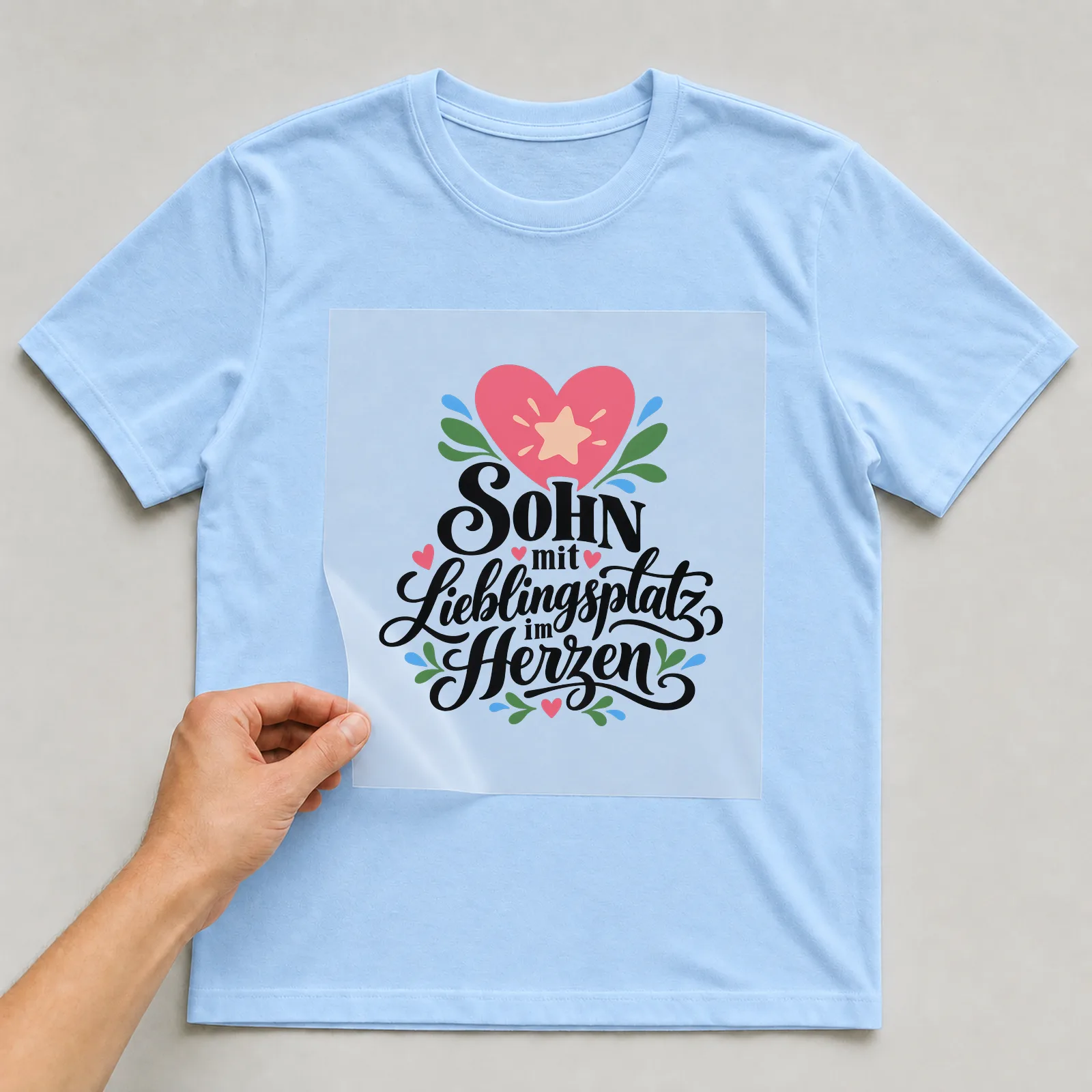 Bügelbild „Sohn mit Lieblingsplatz im Herzen“ auf hellblauem T-Shirt, Folie wird aufgelegt