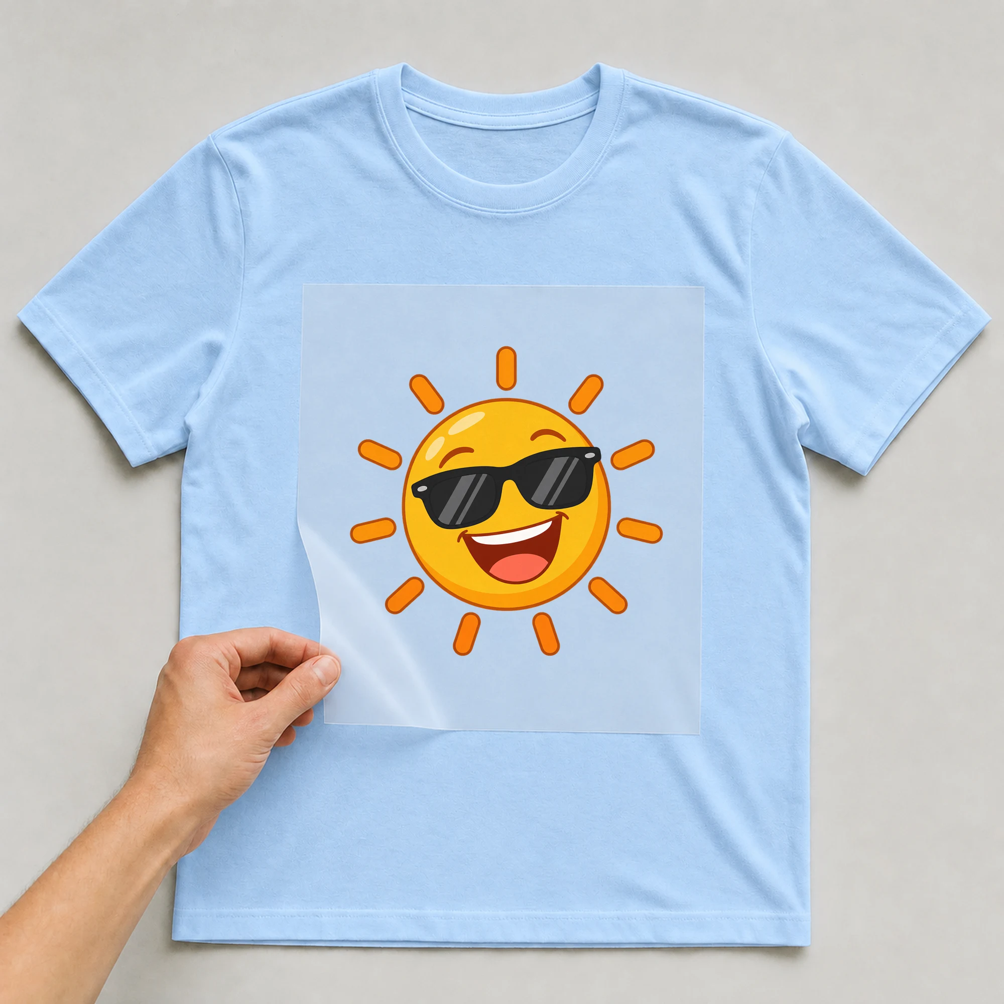 Bügelbild „Sonne mit sonnenbrille motiv ohne text“ auf babyblauem T-Shirt, Folie wird aufgelegt