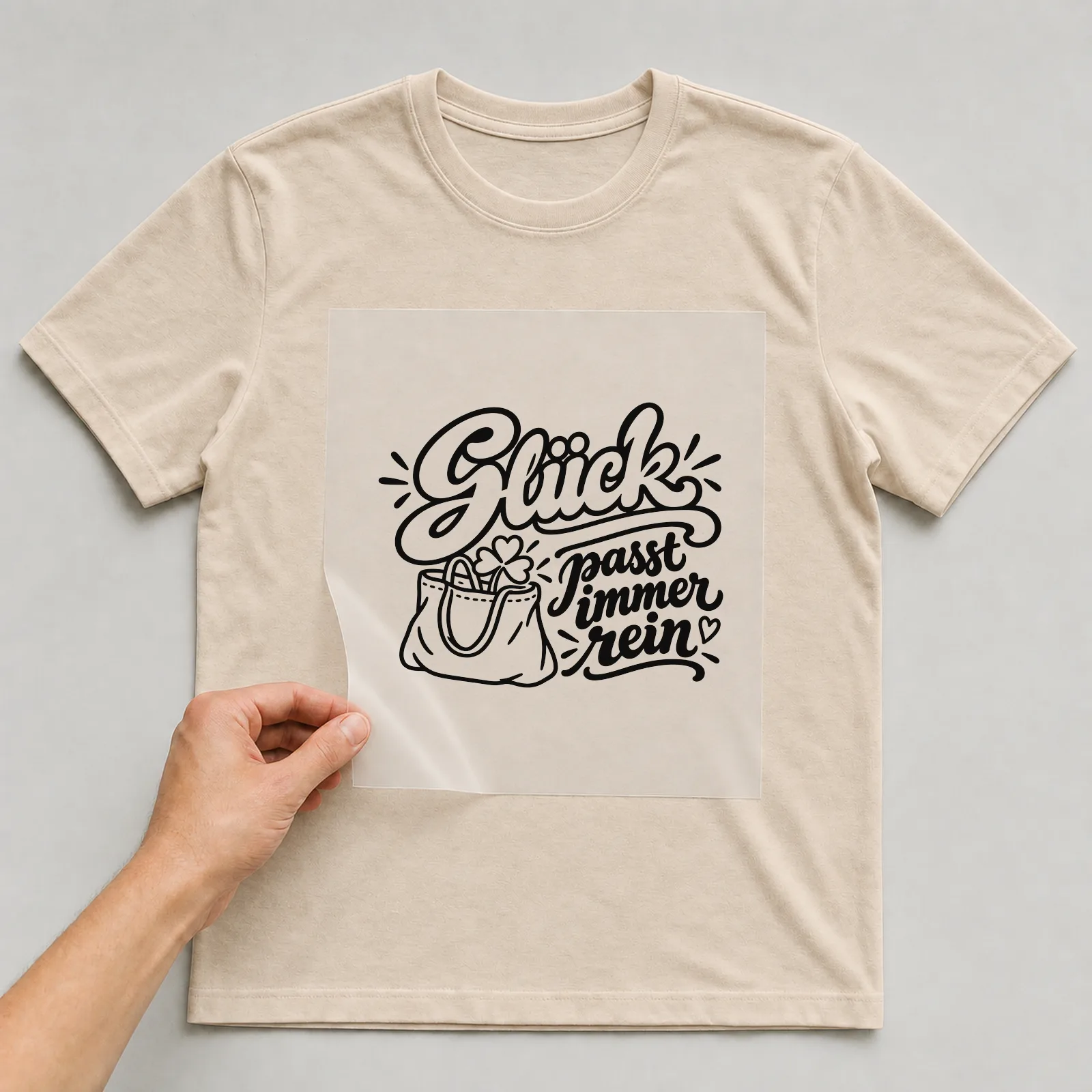 Bügelbild „Glück passt immer rein“ auf beigem T-Shirt, Folie wird aufgelegt