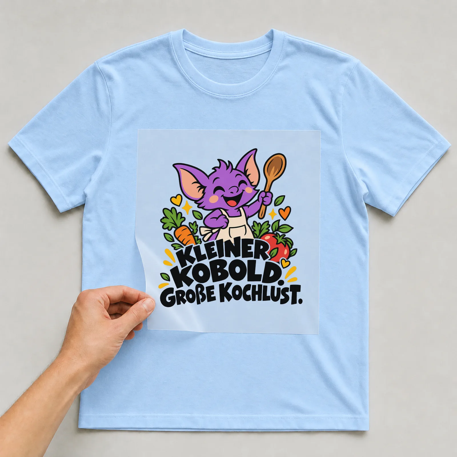 Bügelbild „Kleiner Kobold. Große Kochlust.“ auf hellblauem T-Shirt, Folie wird aufgelegt