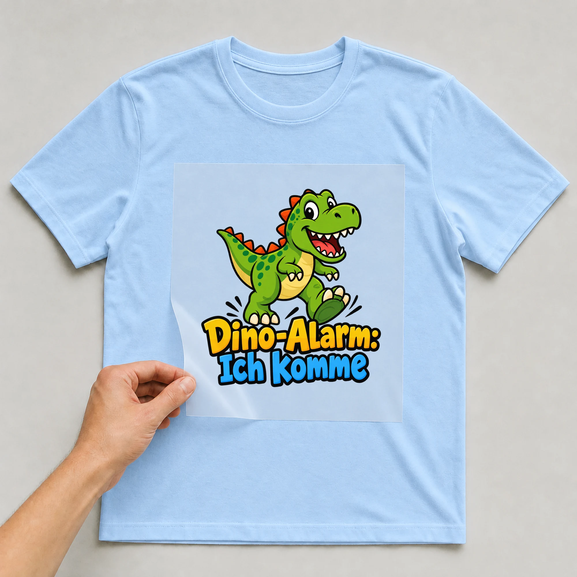 Bügelbild „Dino-Alarm: Ich komme“ auf babyblauem T-Shirt, Folie wird aufgelegt