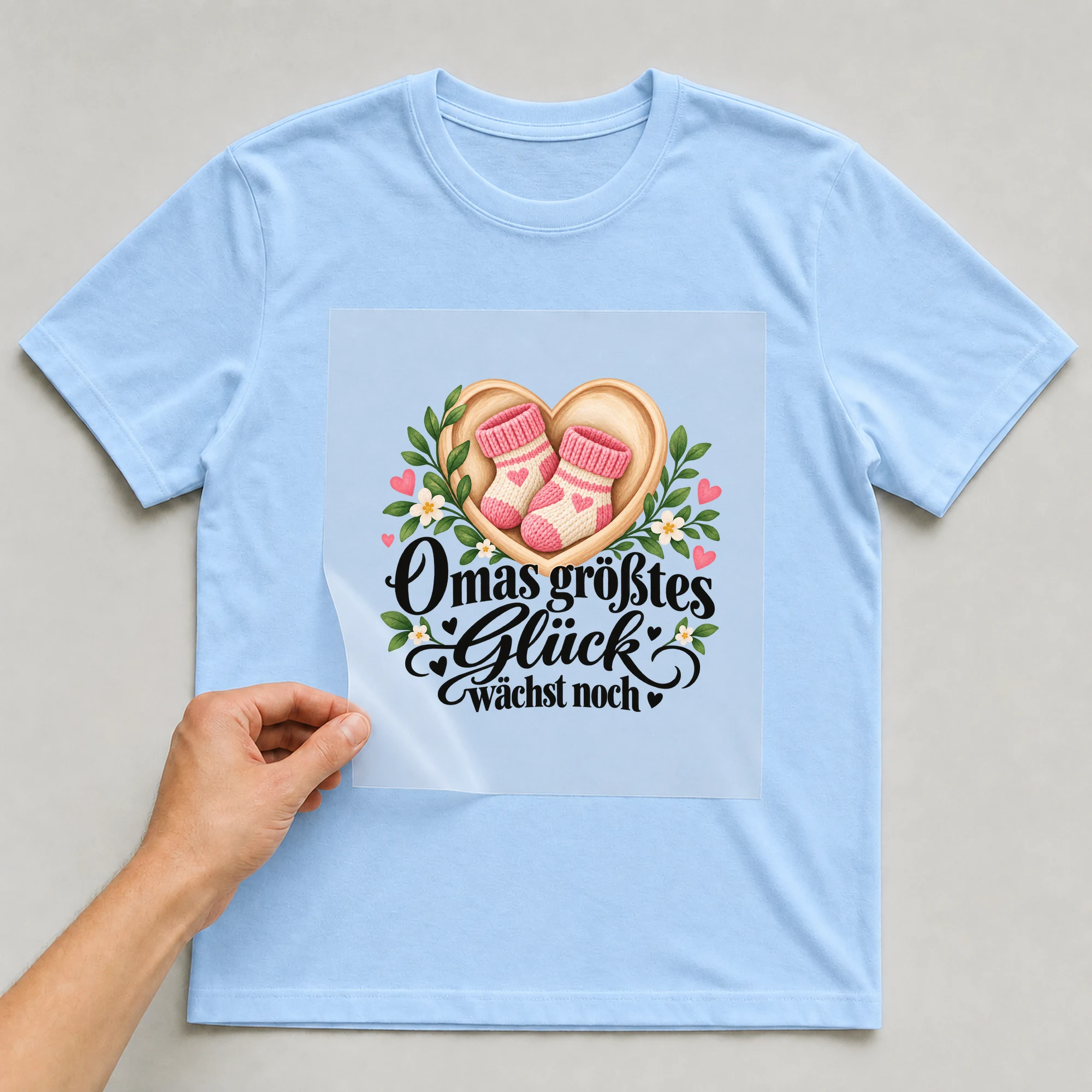 Bügelbild „Omas größtes Glück wächst noch“ auf hellblauem T-Shirt, Folie wird aufgelegt