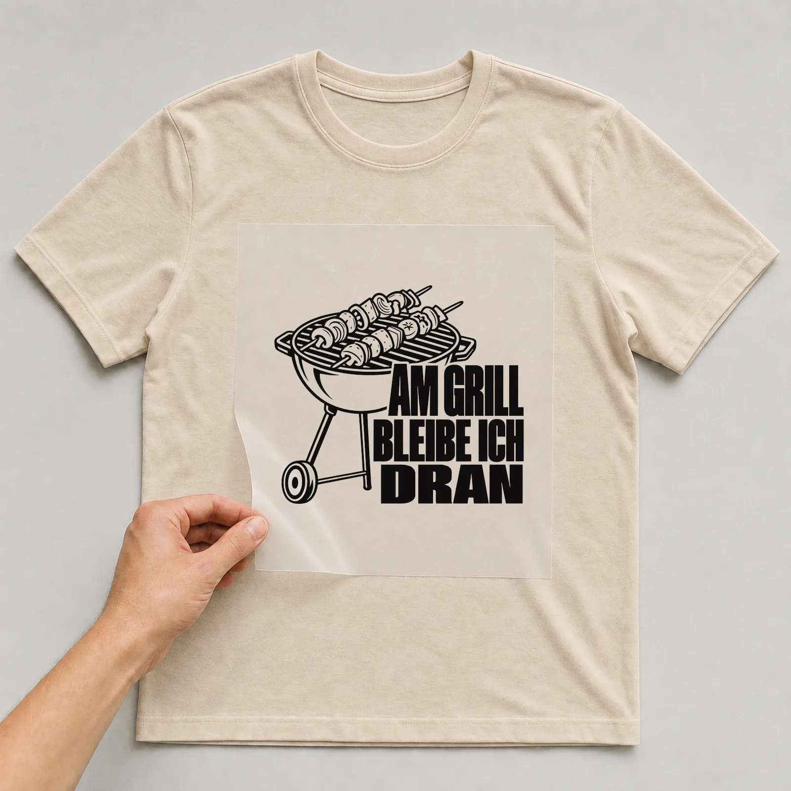 Bügelbild „Am Grill bleibe ich dran“ auf beigem T-Shirt, Folie wird aufgelegt