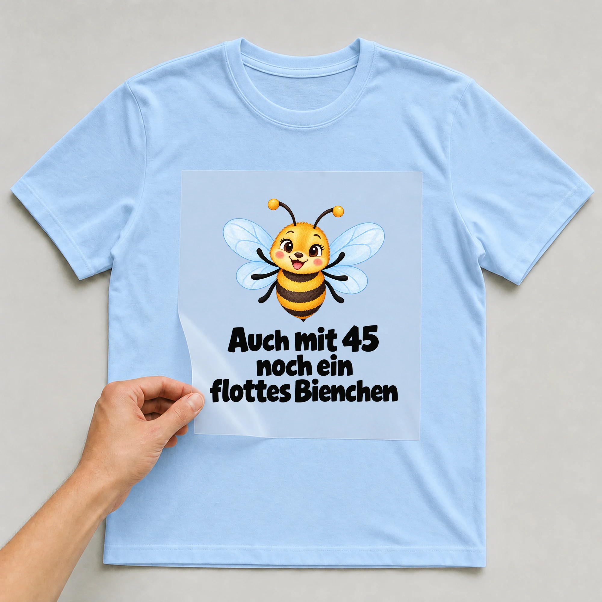 Bügelbild „Auch mit 45 noch ein flottes Bienchen“ auf babyblauem T-Shirt, Folie wird aufgelegt