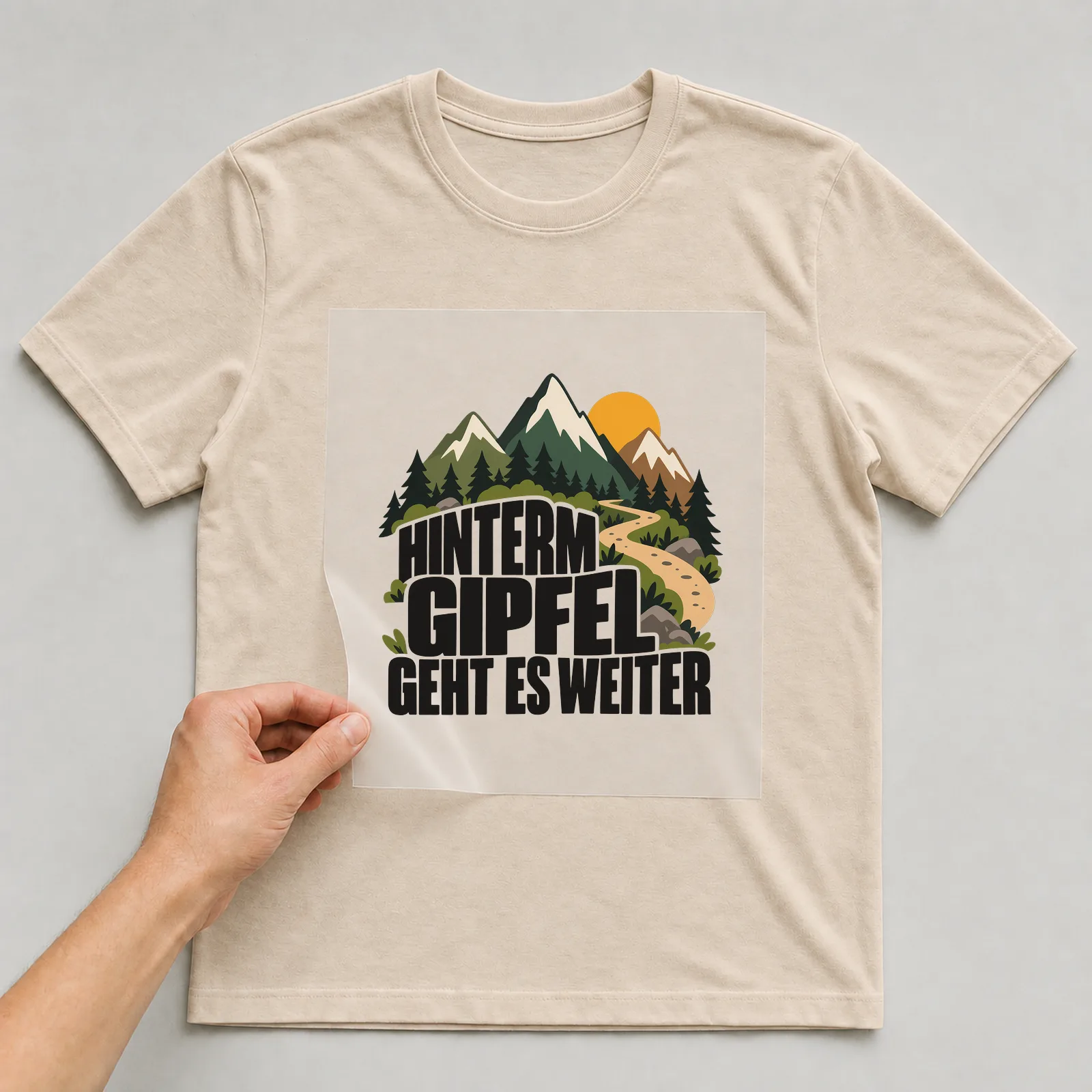 Bügelbild „Hinterm Gipfel geht es weiter“ auf beigem T-Shirt, Folie wird aufgelegt