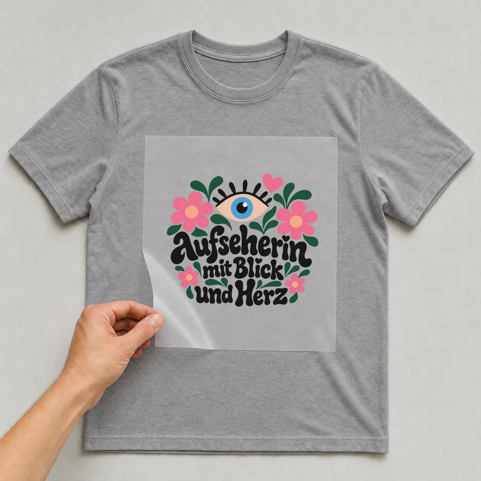 Bügelbild „Aufseherin mit Blick und Herz“ auf grauem T-Shirt, Folie wird aufgelegt