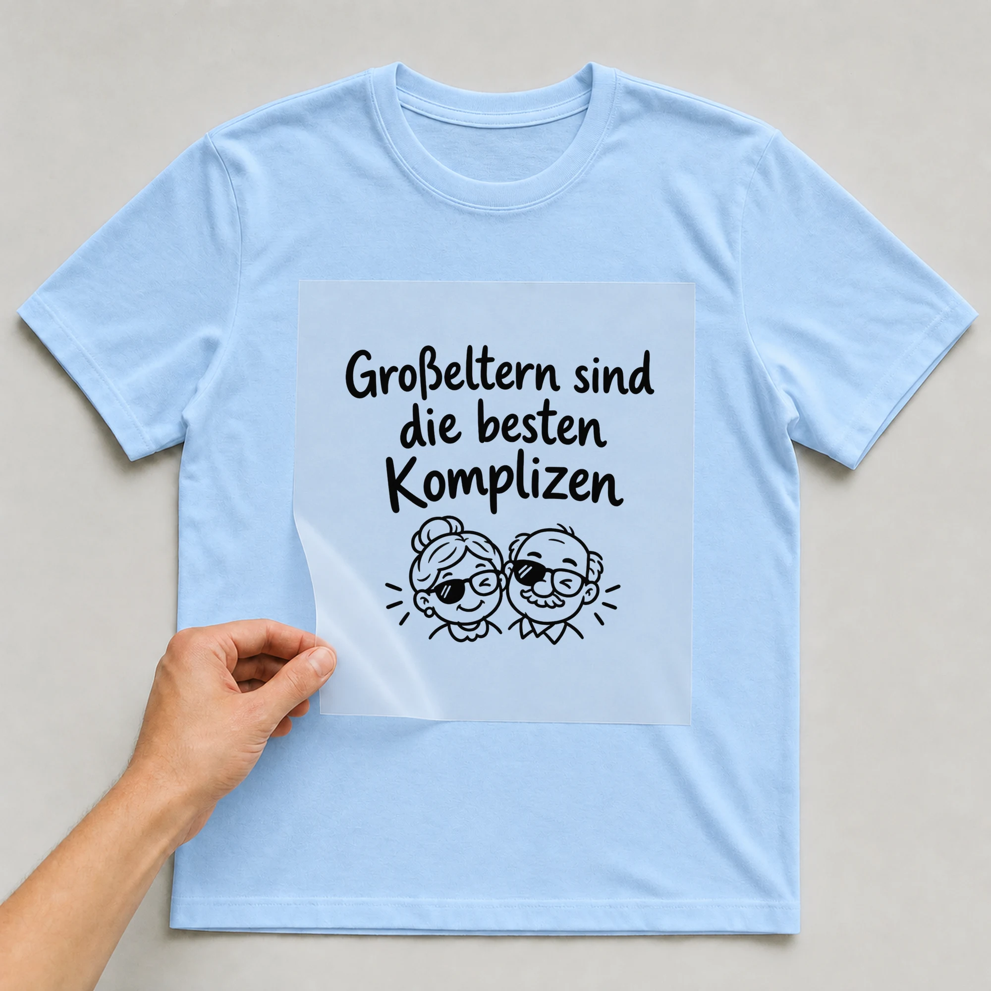 Bügelbild „Großeltern sind die besten Komplizen“ auf babyblauem T-Shirt, Folie wird aufgelegt