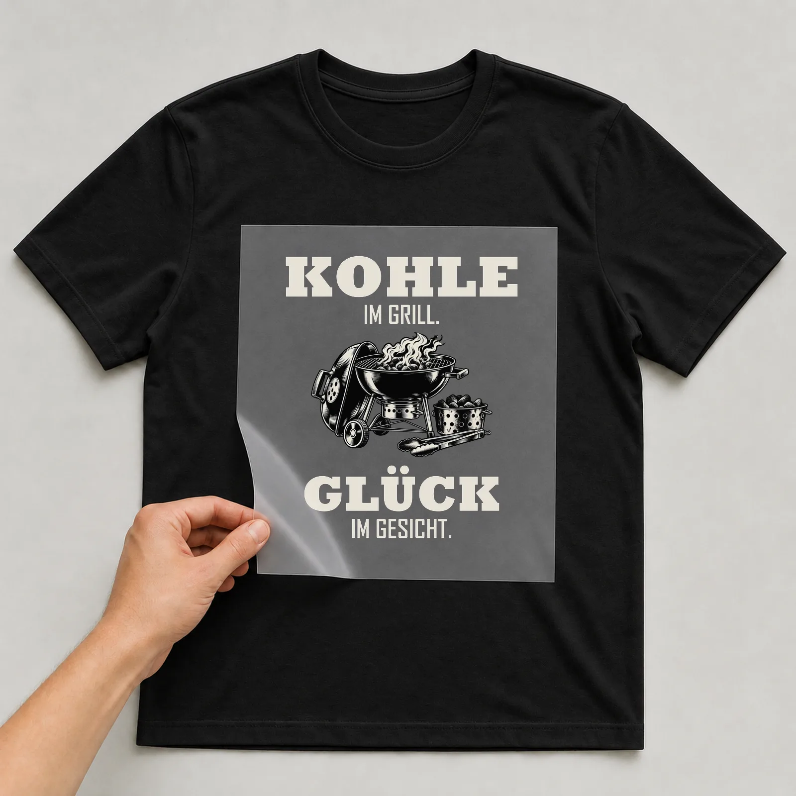 Bügelbild „Kohle im Grill. Glück im Gesicht.“ auf schwarzem T-Shirt, Folie wird aufgelegt