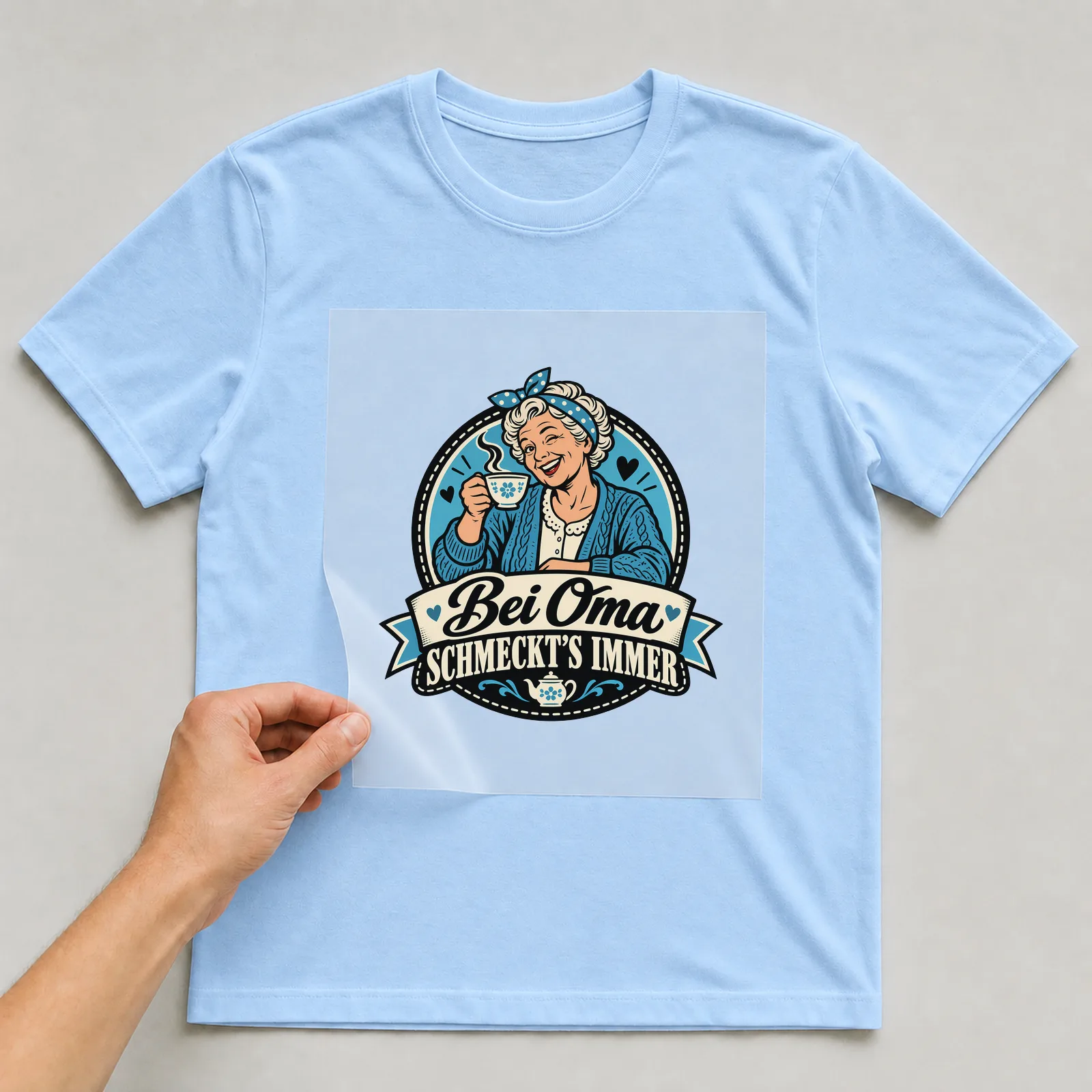 Bügelbild „Bei Oma schmeckt’s immer“ auf hellblauem T-Shirt, Folie wird aufgelegt