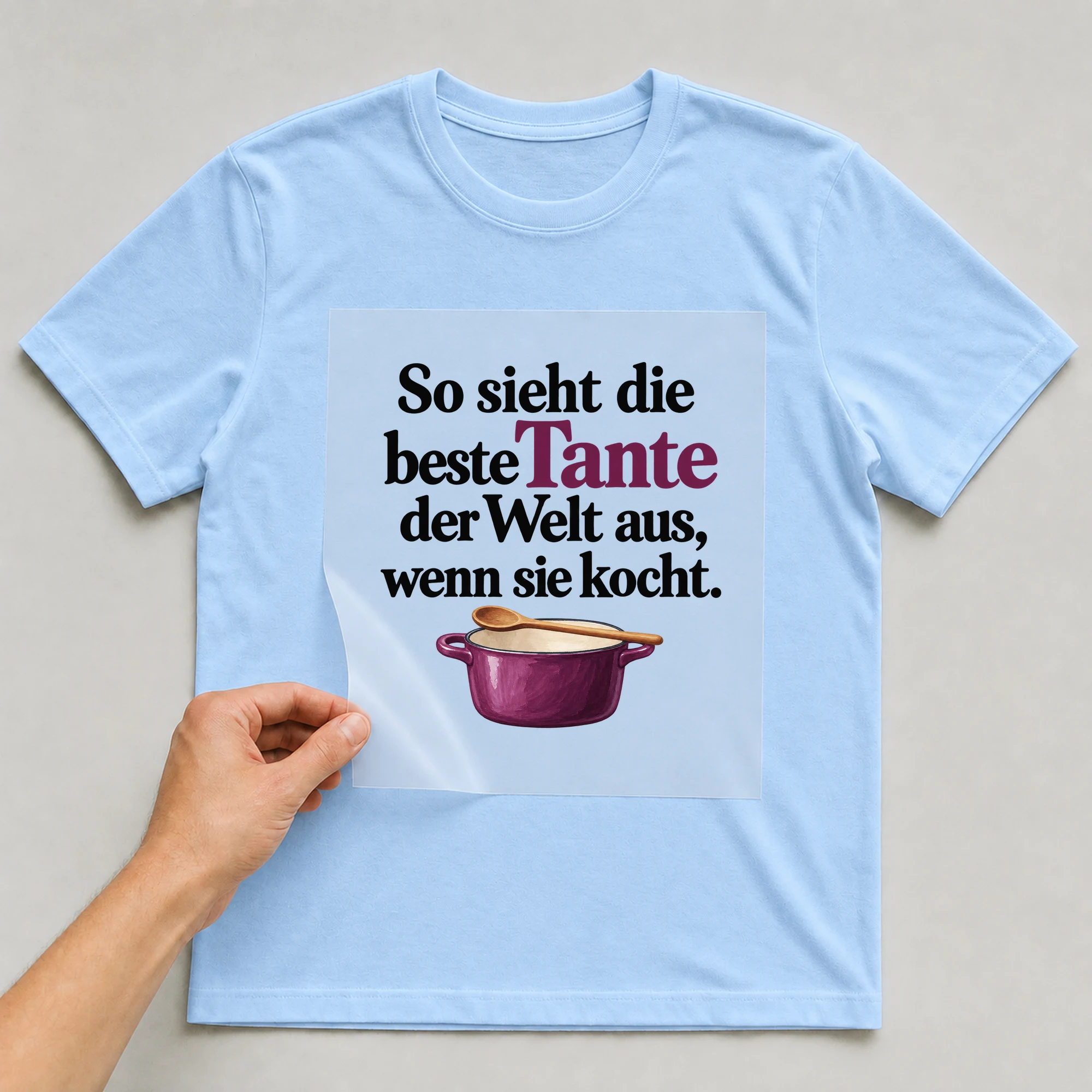 Bügelbild „So sieht die beste Tante der Welt aus, wenn sie kocht.“ auf babyblauem T-Shirt, Folie wird aufgelegt