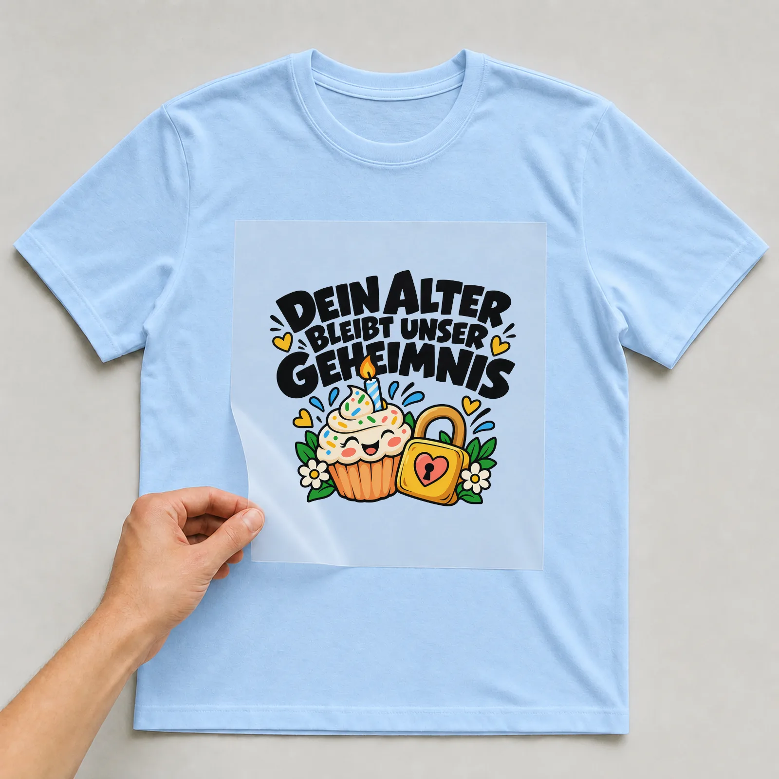 Bügelbild „Dein Alter bleibt unser Geheimnis“ auf hellblauem T-Shirt, Folie wird aufgelegt