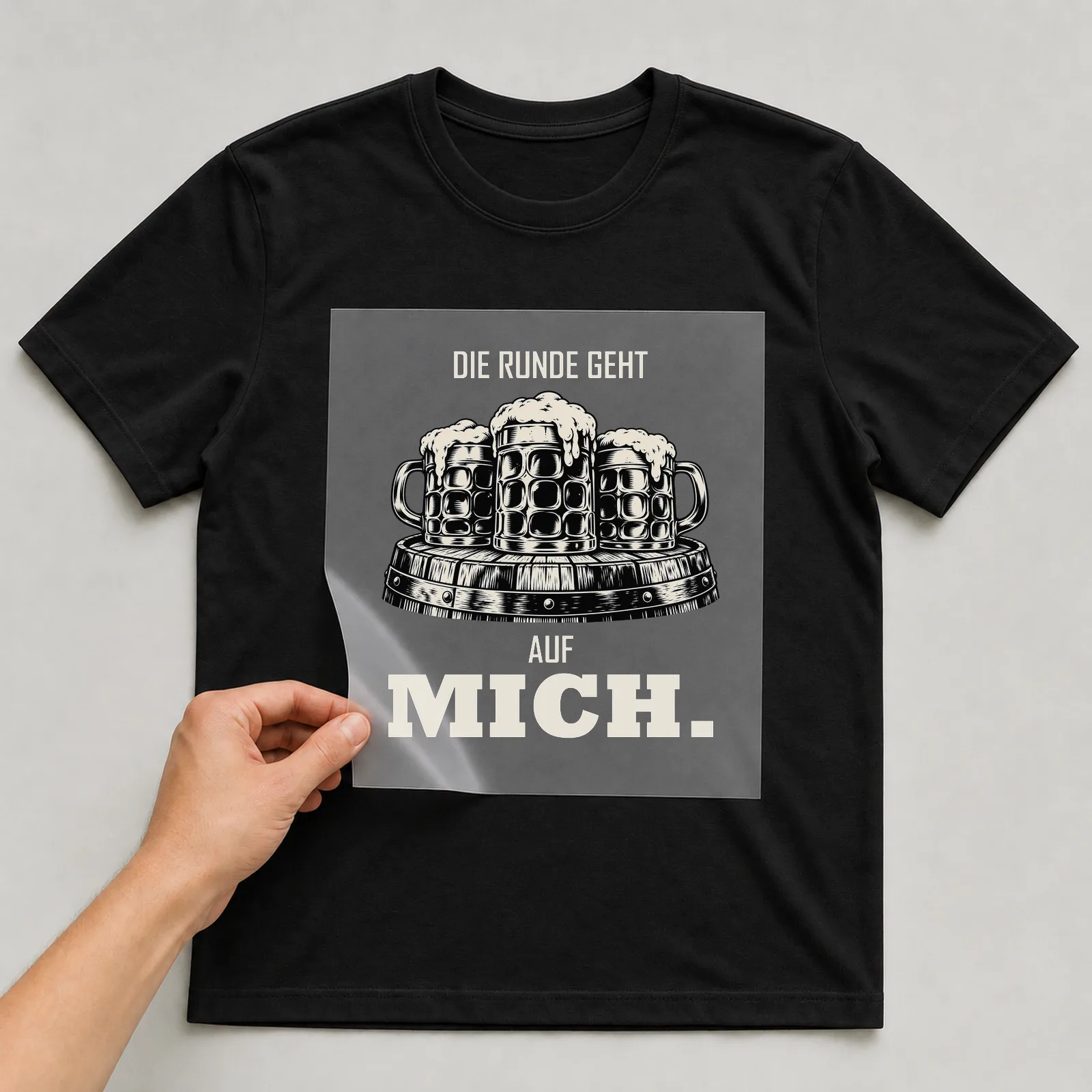 Bügelbild „Die Runde geht auf mich“ auf schwarzem T-Shirt, Folie wird aufgelegt