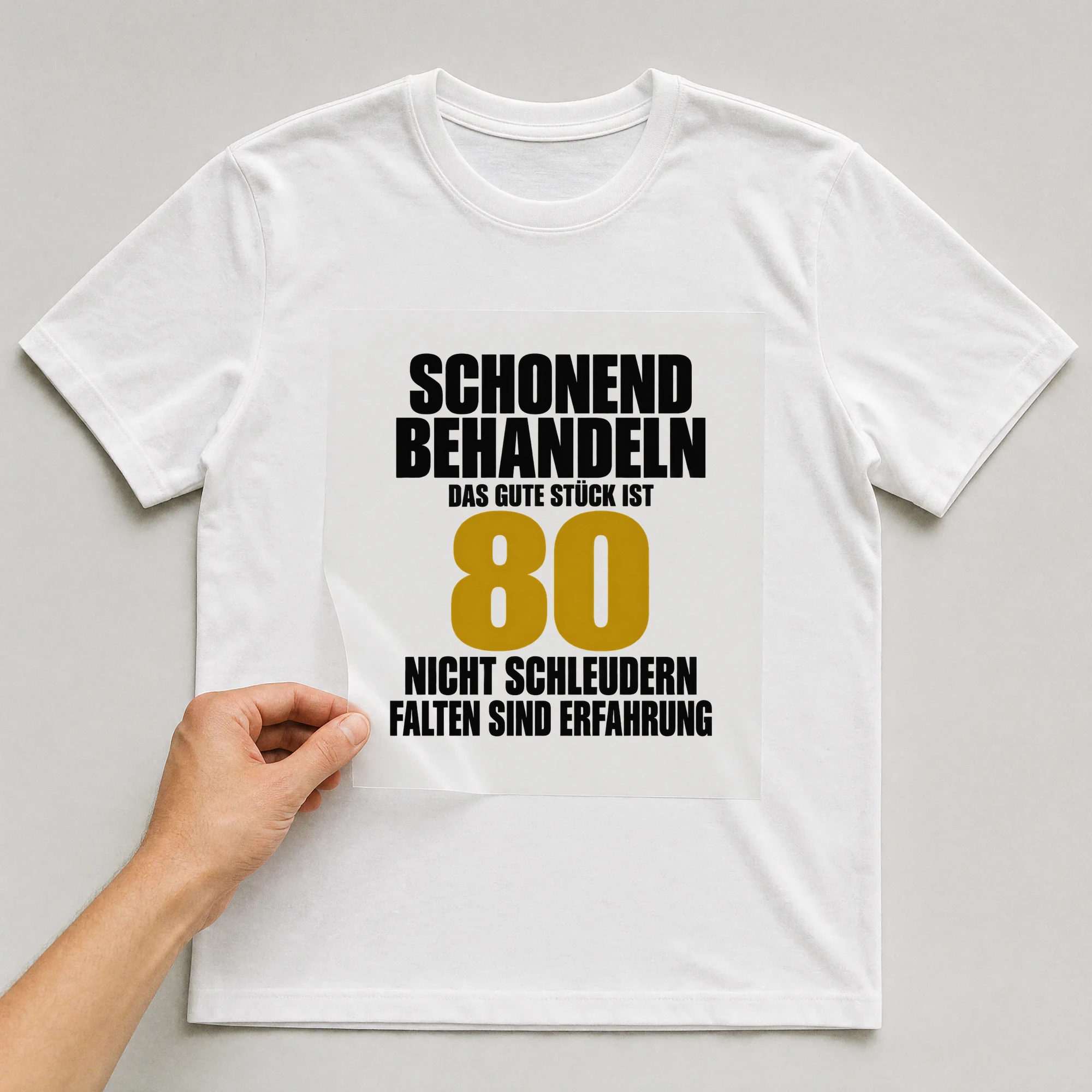 Bügelbild „Schonend behandeln: Das gute Stück ist 80.“ auf weißem T-Shirt, Folie wird aufgelegt