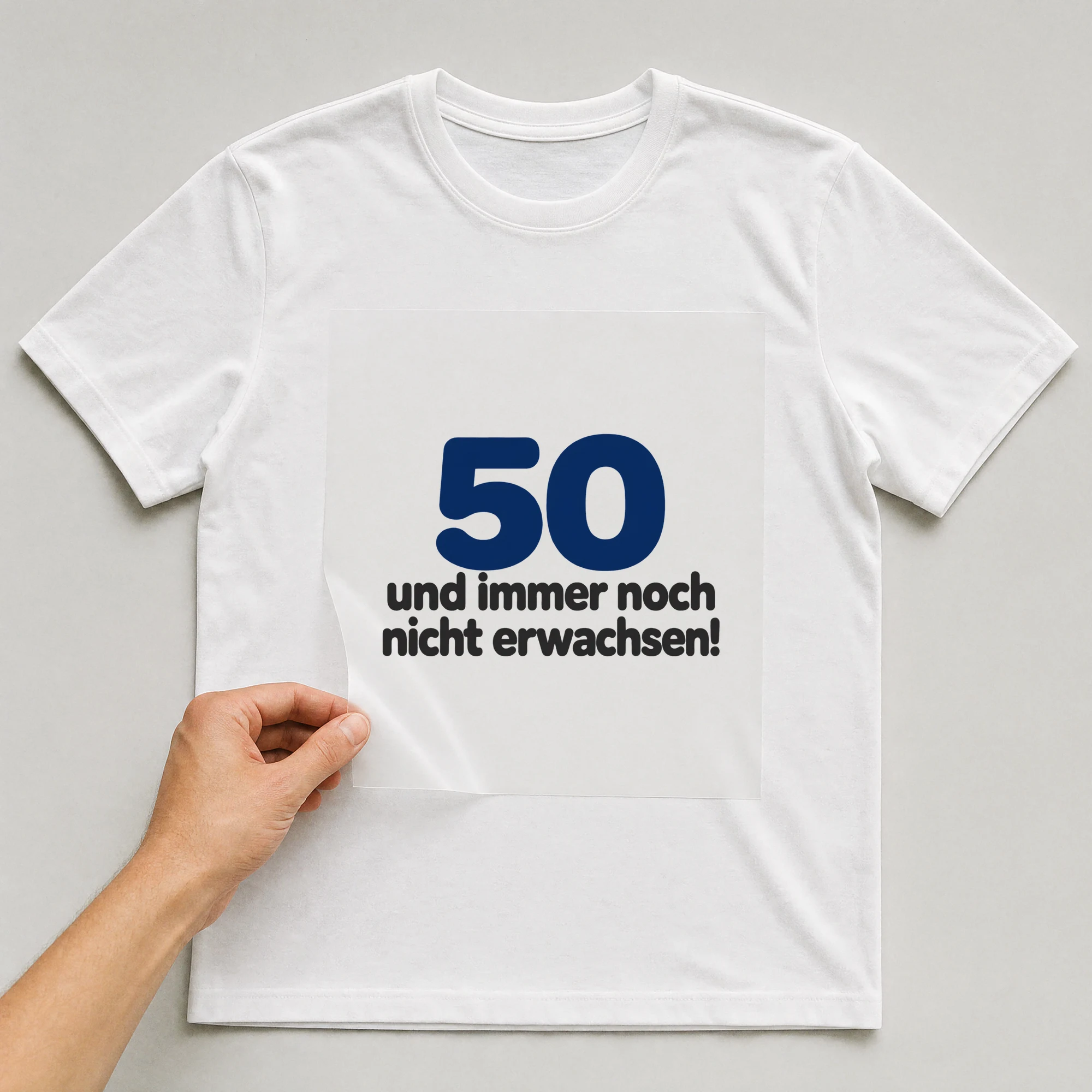 Bügelbild „50 und immer noch nicht erwachsen!“ auf weißem T-Shirt, Folie wird aufgelegt