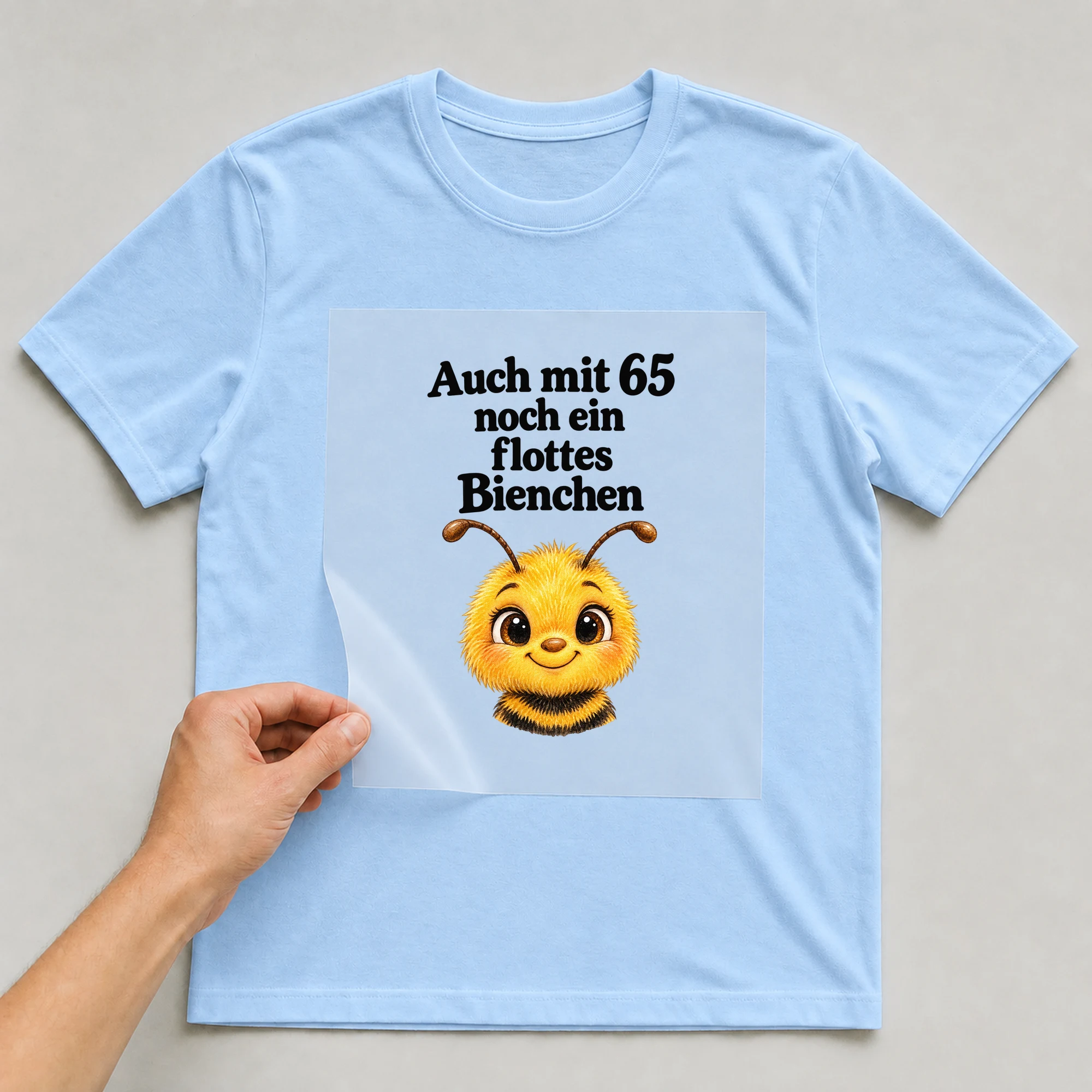 Bügelbild „Auch mit 65 noch ein flottes Bienchen“ auf babyblauem T-Shirt, Folie wird aufgelegt