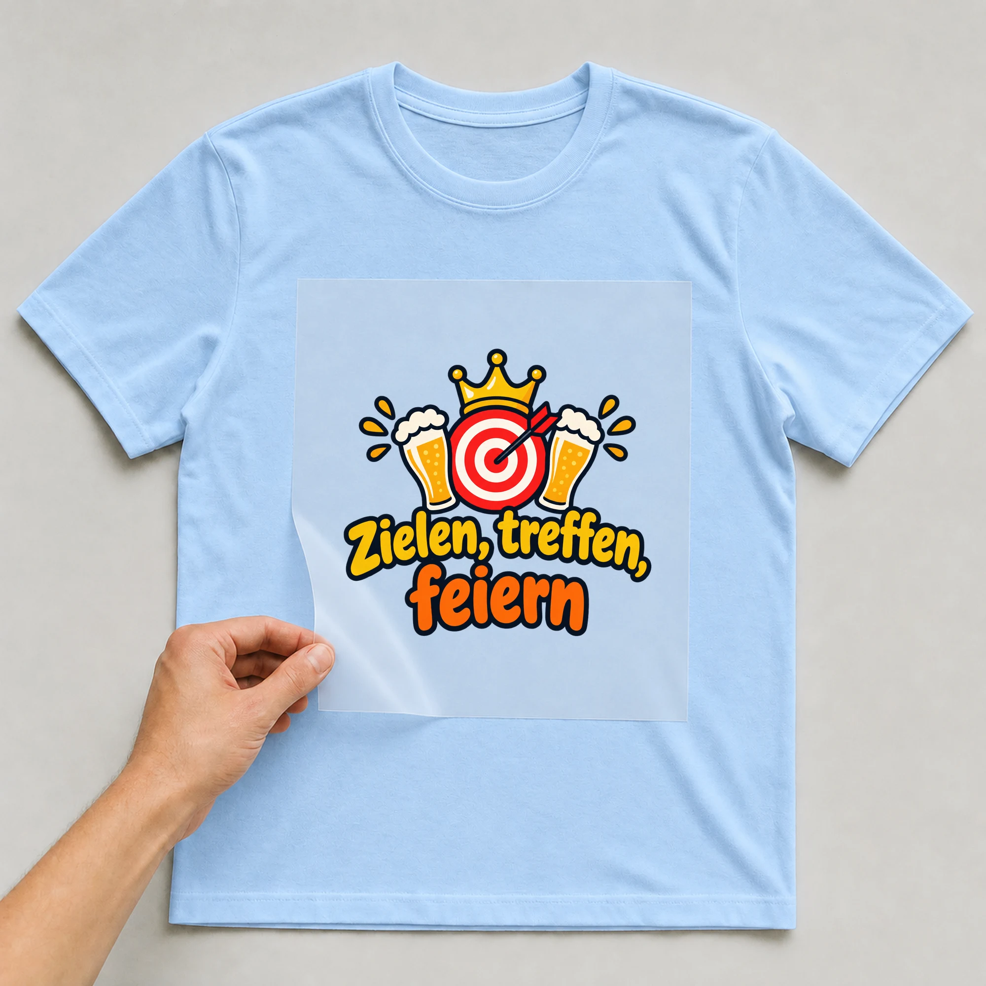 Bügelbild „Zielen, treffen, feiern“ auf babyblauem T-Shirt, Folie wird aufgelegt